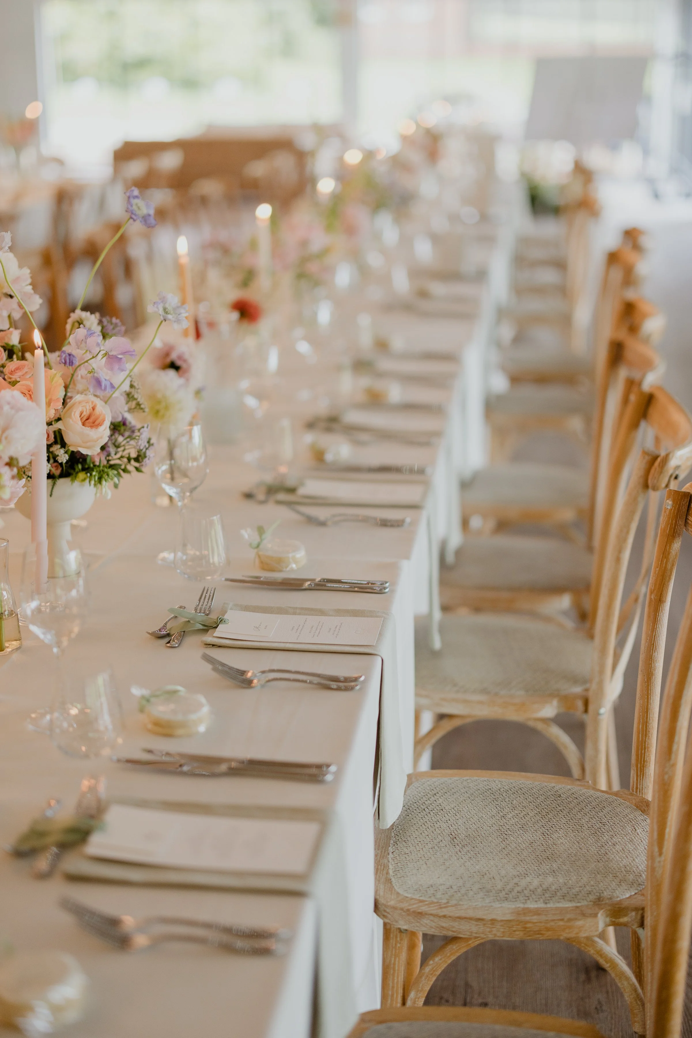 A tablescape with a centerpiece of pink, white, and purple flowers in a white vase, surrounded by smaller vases, candles, and place settings with silverware, napkins, and menus in a bright, well-lit room with large windows and wooden chairs.