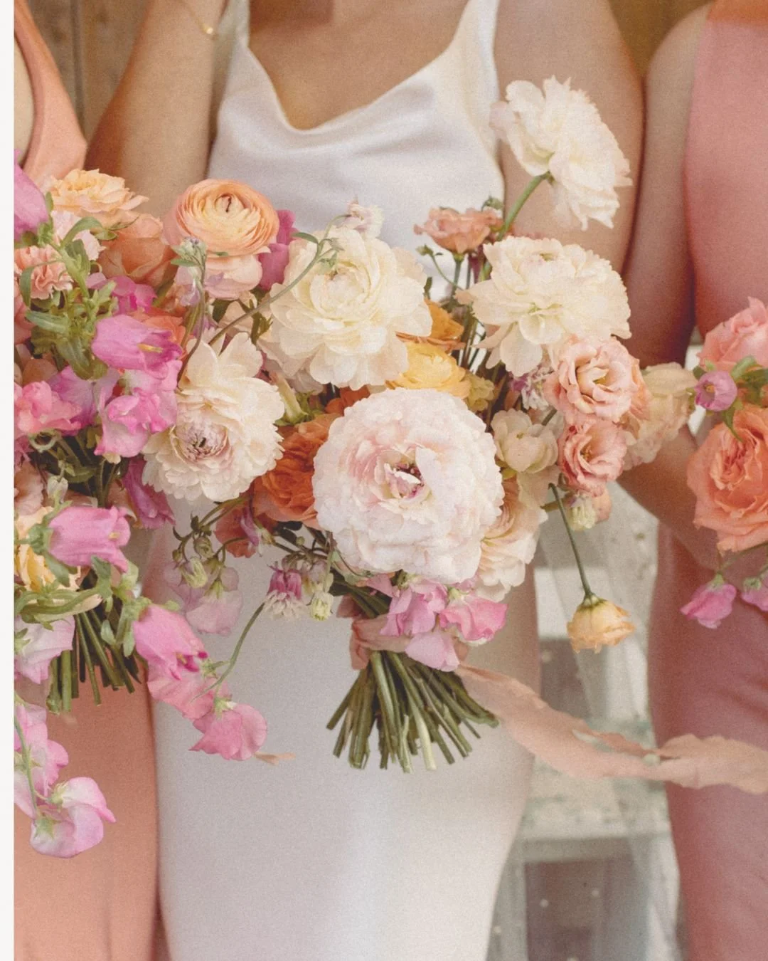 Close-up of people holding a bouquet of flowers with pink, white, and peach blooms, including roses and ranunculus, in front of pastel-colored dresses.