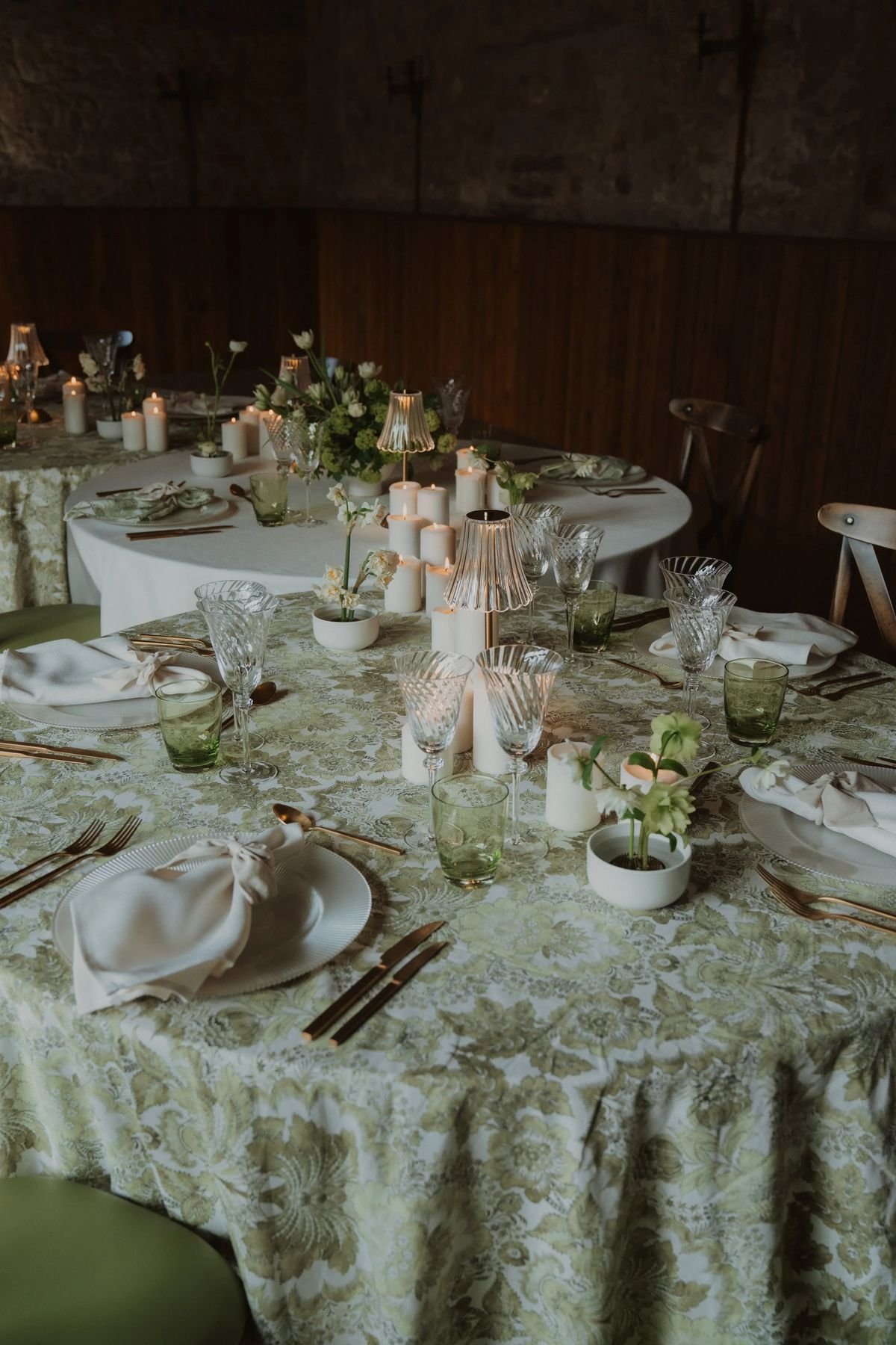 Elegant dinner table setup with floral tablecloths, white plates, glassware, candles, and floral centerpieces.