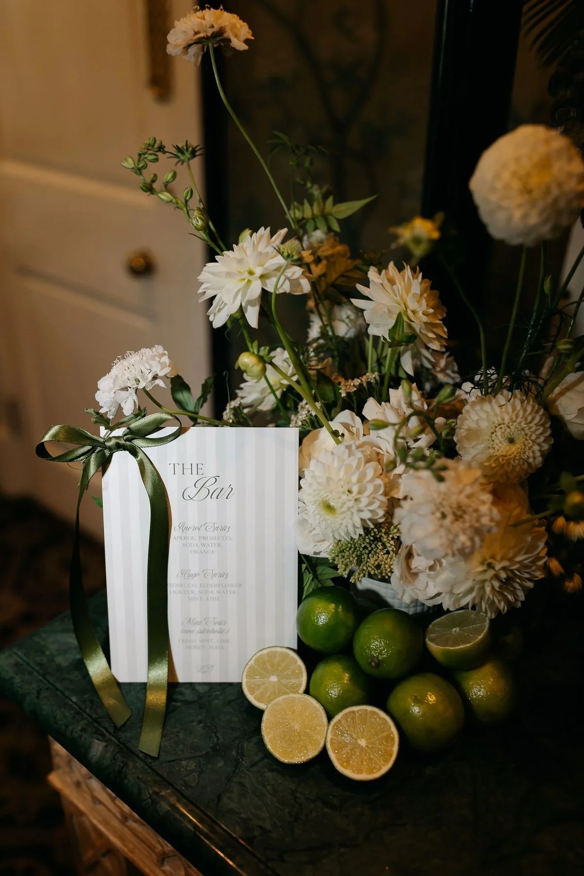 A floral arrangement with white and cream-colored flowers, green foliage, and limes, including some cut in half showing their yellow interior, on a dark green marble table. A white striped menu card with elegant script is also part of the display.