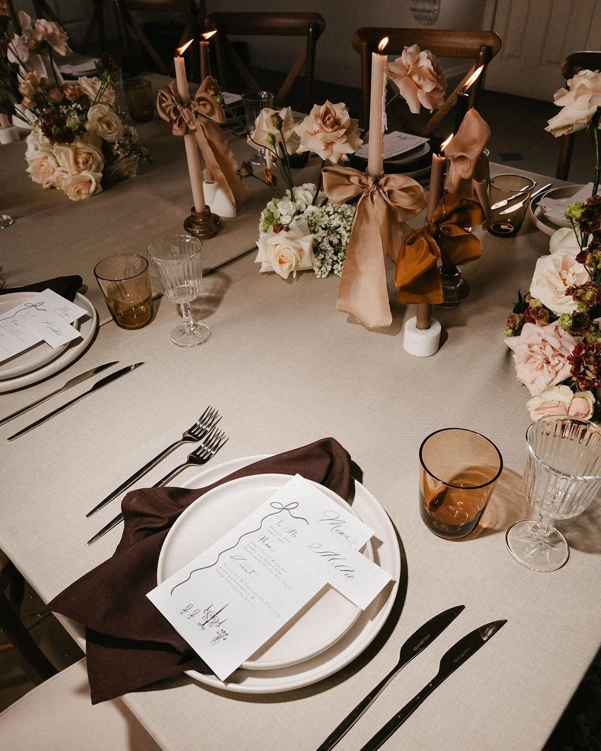 Elegant dining table setup with white dinner plates, brown napkins, a printed menu, glassware, and silverware. Decor includes pink candles with bows, and floral centerpieces with roses and greenery on a beige tablecloth.