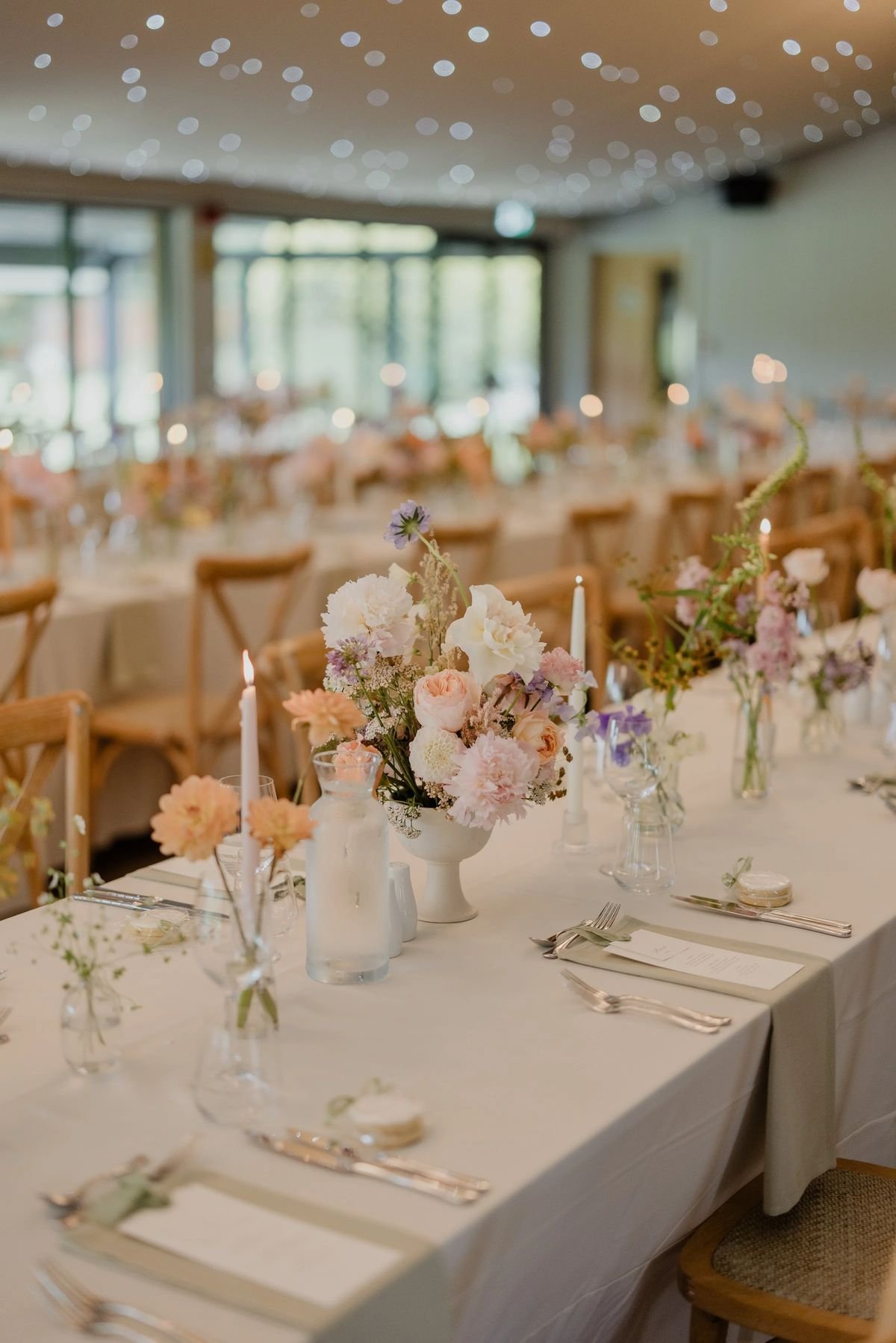A tablescape with a centerpiece of pink, white, and purple flowers in a white vase, surrounded by smaller vases, candles, and place settings with silverware, napkins, and menus in a bright, well-lit room with large windows and wooden chairs.