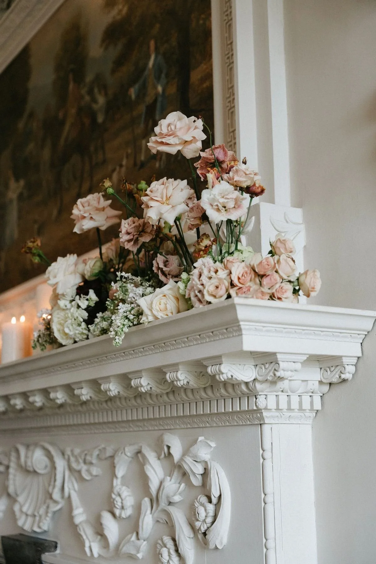 A bouquet of dried pink and white roses and white flowers on a white fireplace mantel with ornate carvings, with a painting of two people riding horses in the background.