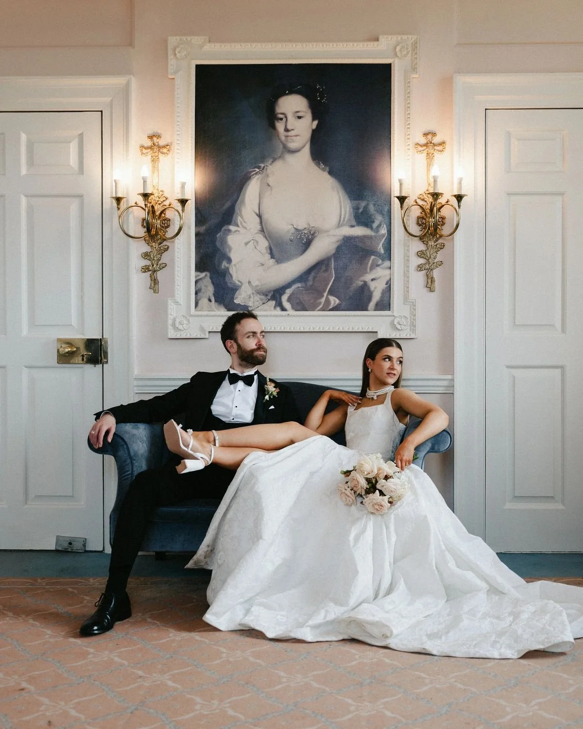A bride and groom sitting on a blue sofa in a formal room with white paneled walls and a large portrait of a woman in historical attire hanging on the wall behind them. The bride is reclining with her legs crossed, holding a bouquet of flowers, wearing a white wedding gown and jewelry. The groom sits with his arm resting on the back of the sofa, dressed in a tuxedo with a bow tie, with a boutonnière on his lapel, and has a beard. The room is decorated with ornate lighting fixtures and framed with decorative molding.