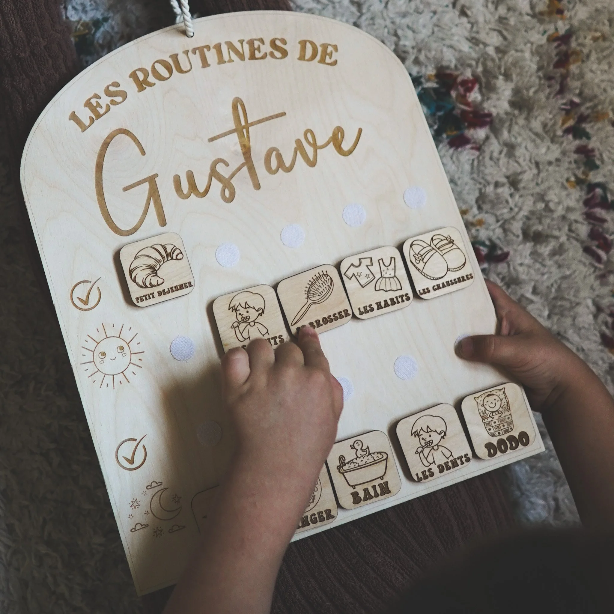Tableau en bois intitulé 'Les Routines de Gustave' avec des icônes représentant des activités quotidiennes comme le petit-déjeuner, la toilette, l'habillement et le coucher. Une main d'enfant pointe vers une icône. Le tableau est posé sur un tapis.