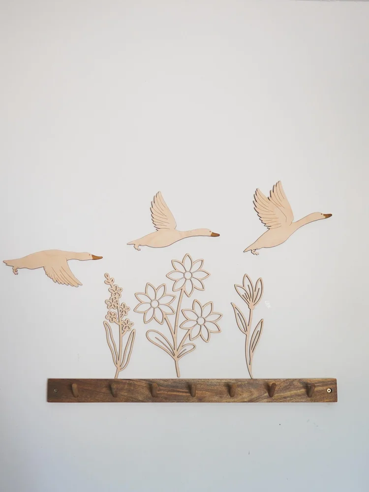 Trois oies en vol et trois fleurs en bois naturel fixées sur un mur blanc au-dessus d'une patère