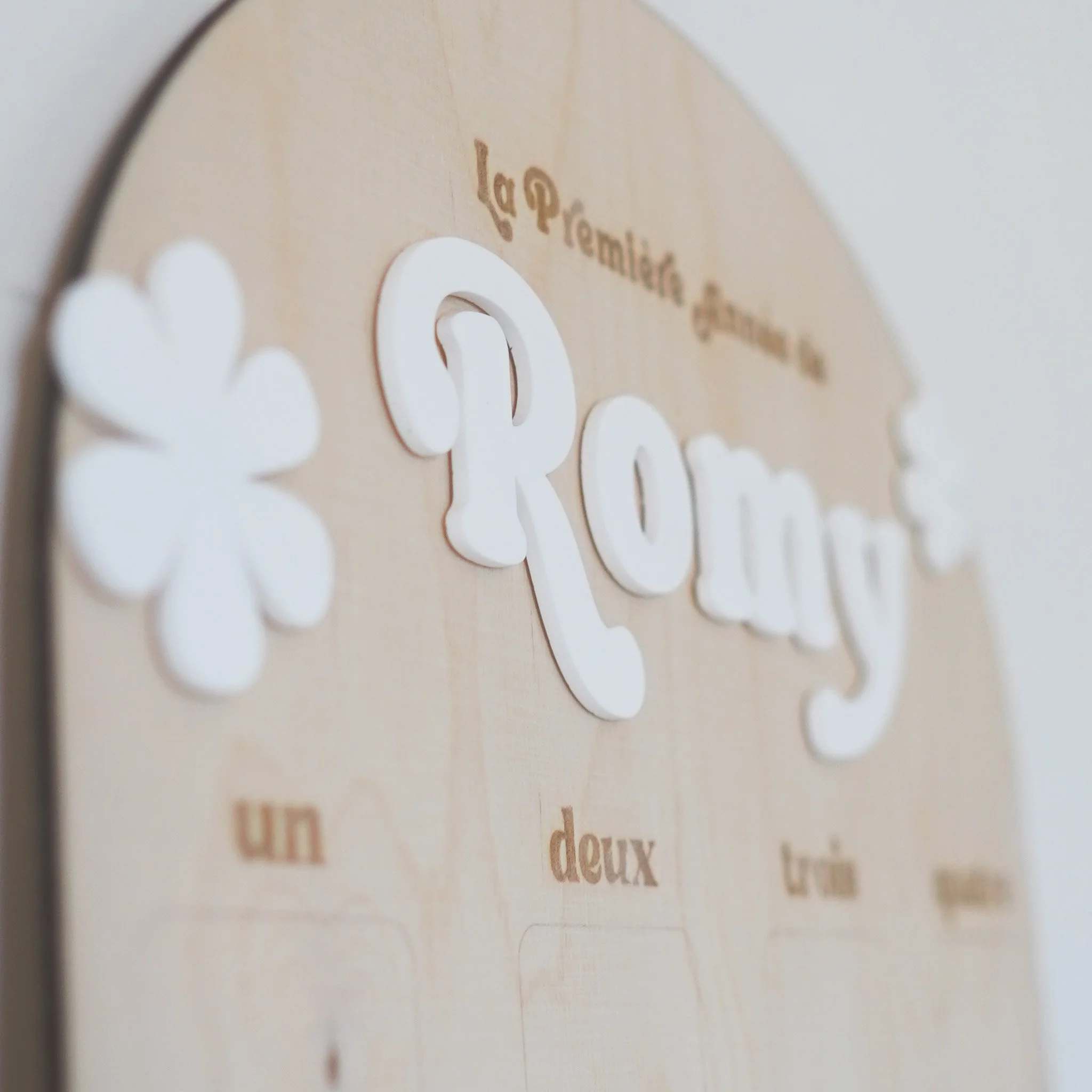 Plaque en bois avec décoration florale et inscription "La Première Année de Romy" en lettres blanches, accompagnée des mots "un", "deux", "trois", "quatre" gravés en dessous.
