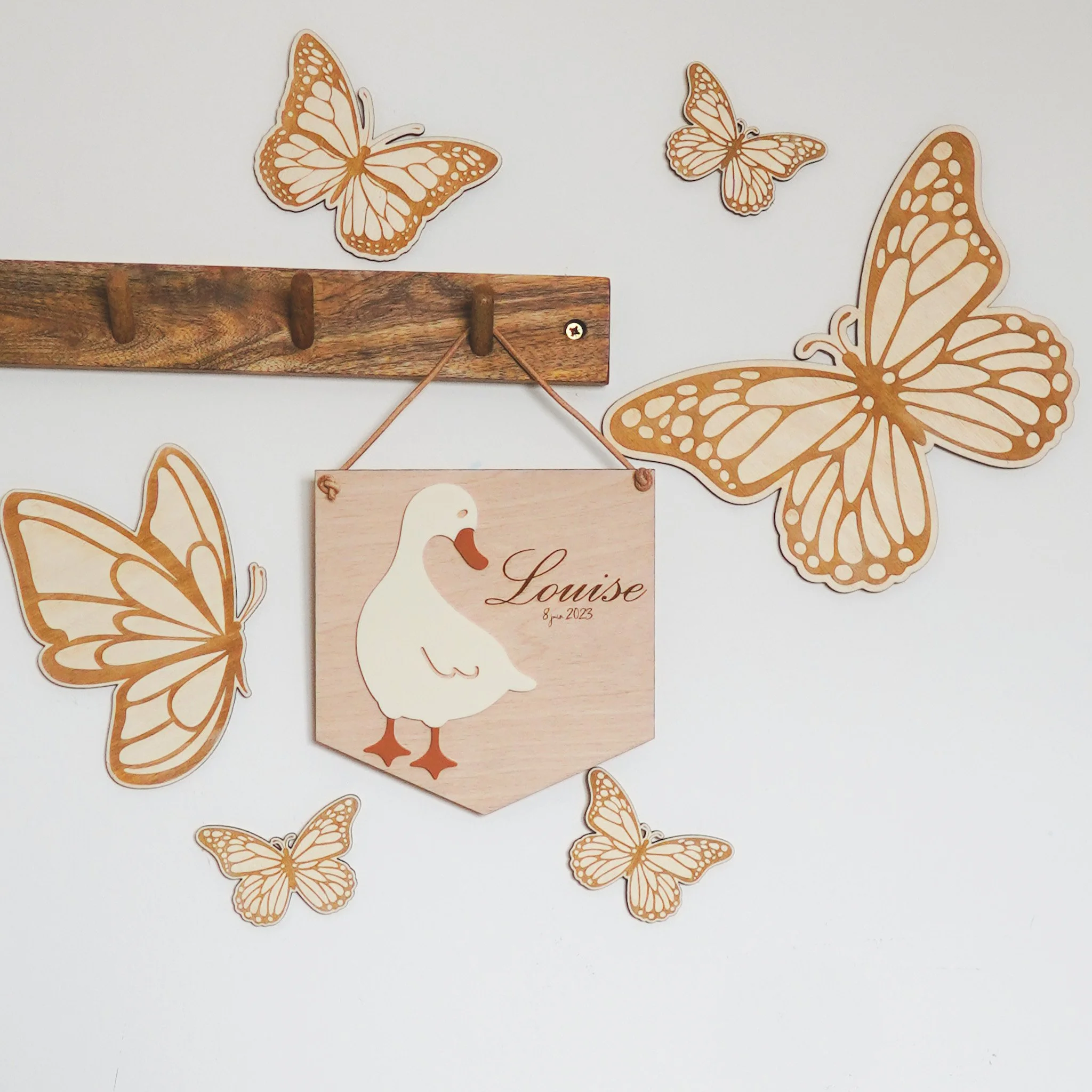 Illustration murale avec des papillons en bois et une planche avec l'image d'un canard et le mot "Louise". Une étagère en bois est également visible au-dessus.