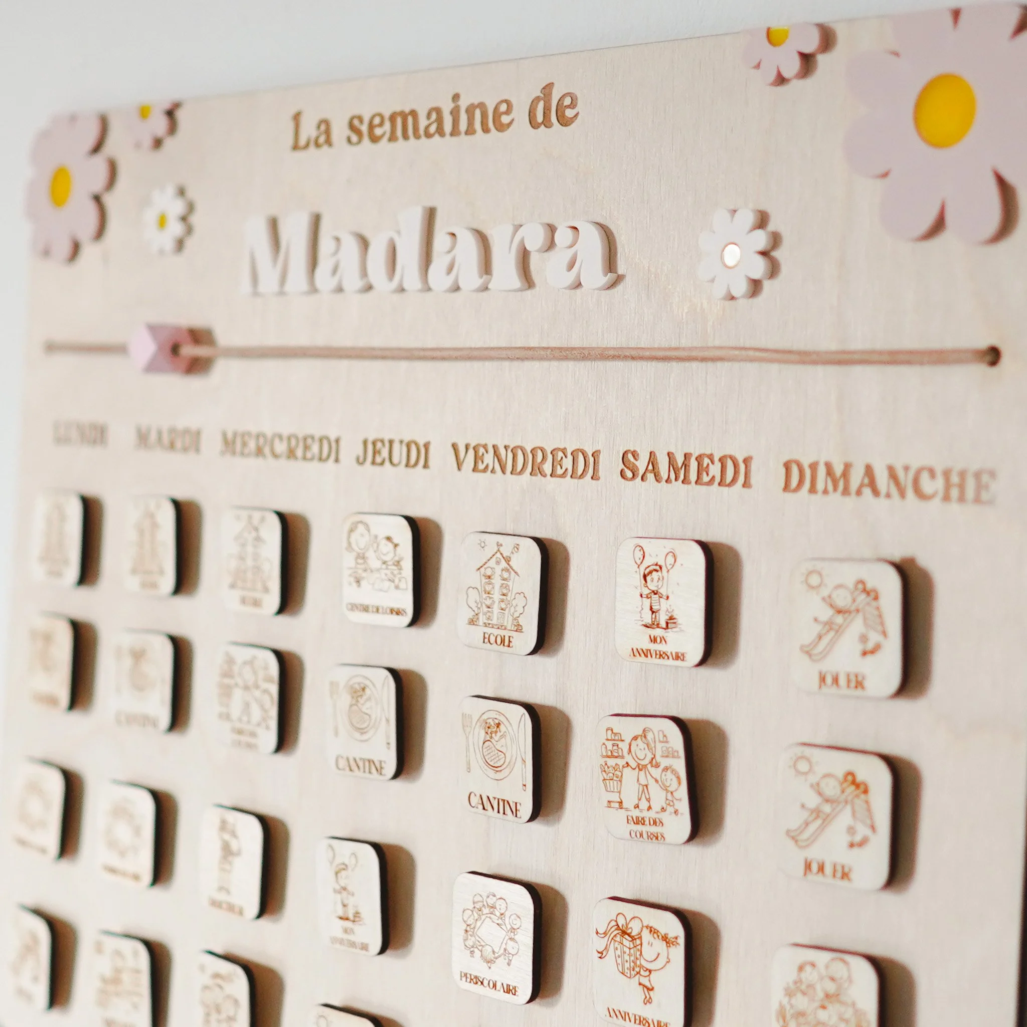 Calendrier hebdomadaire en bois avec des activités pour les jours de la semaine, décoré de fleurs. "La semaine de Madara" est écrit en haut. Chaque jour a plusieurs cases illustrant diverses activités comme l'école, anniversaire, et jouer.