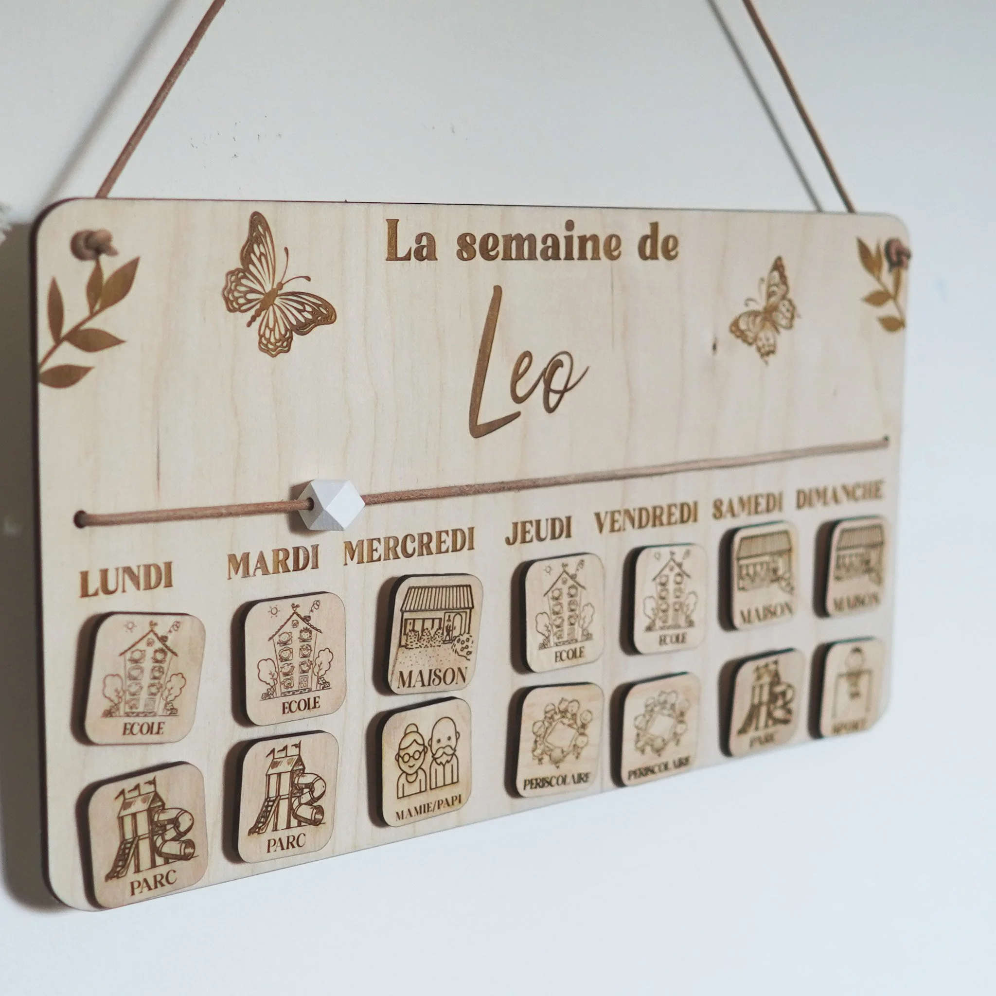 Tableau en bois personnalisable pour enfants montrant l'emploi du temps de la semaine de "Leo". Les jours de la semaine sont écrits avec des illustrations gravées représentant des activités comme école, maison, parc et mamie/papi. Il est suspendu et décoré de motifs de papillons et feuilles.
