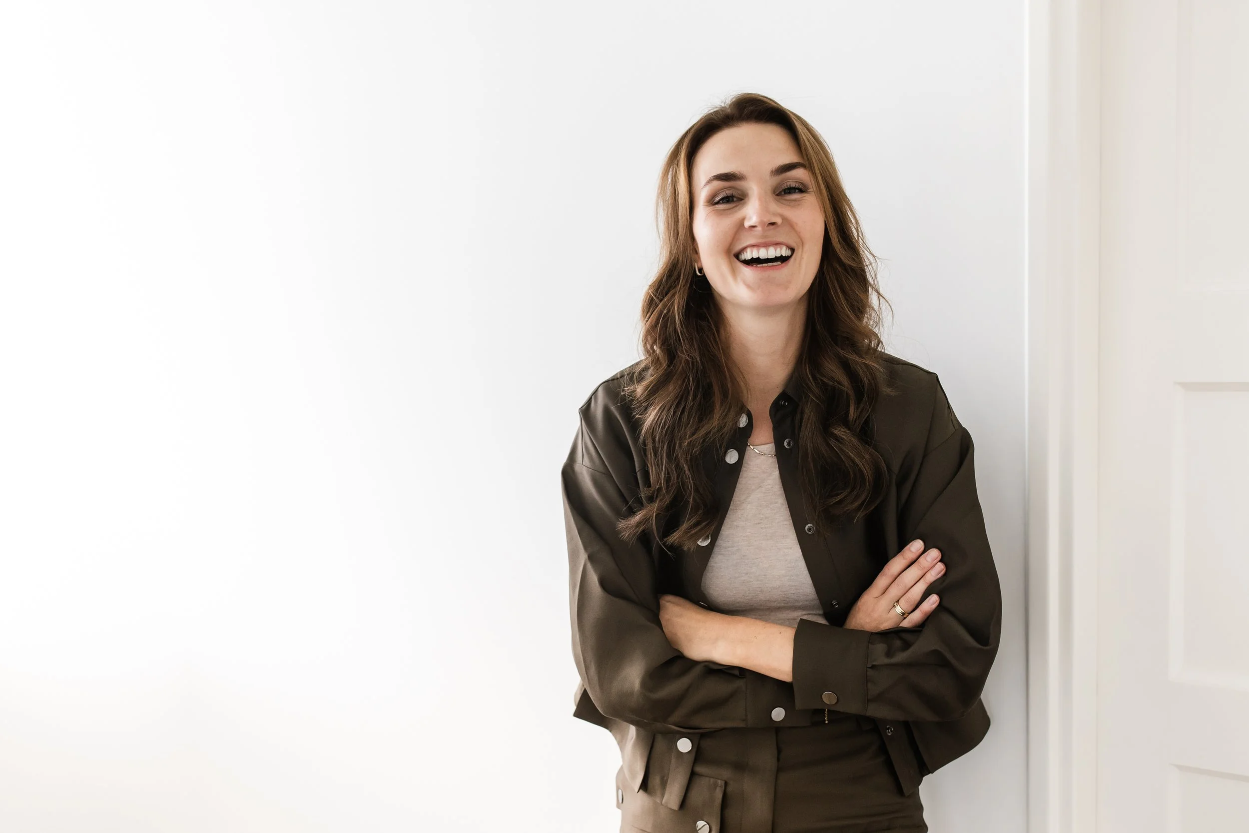 Lachende vrouw met bruin haar, gekleed in een grijze top en een donker groene jas, staand tegen een witte muur.