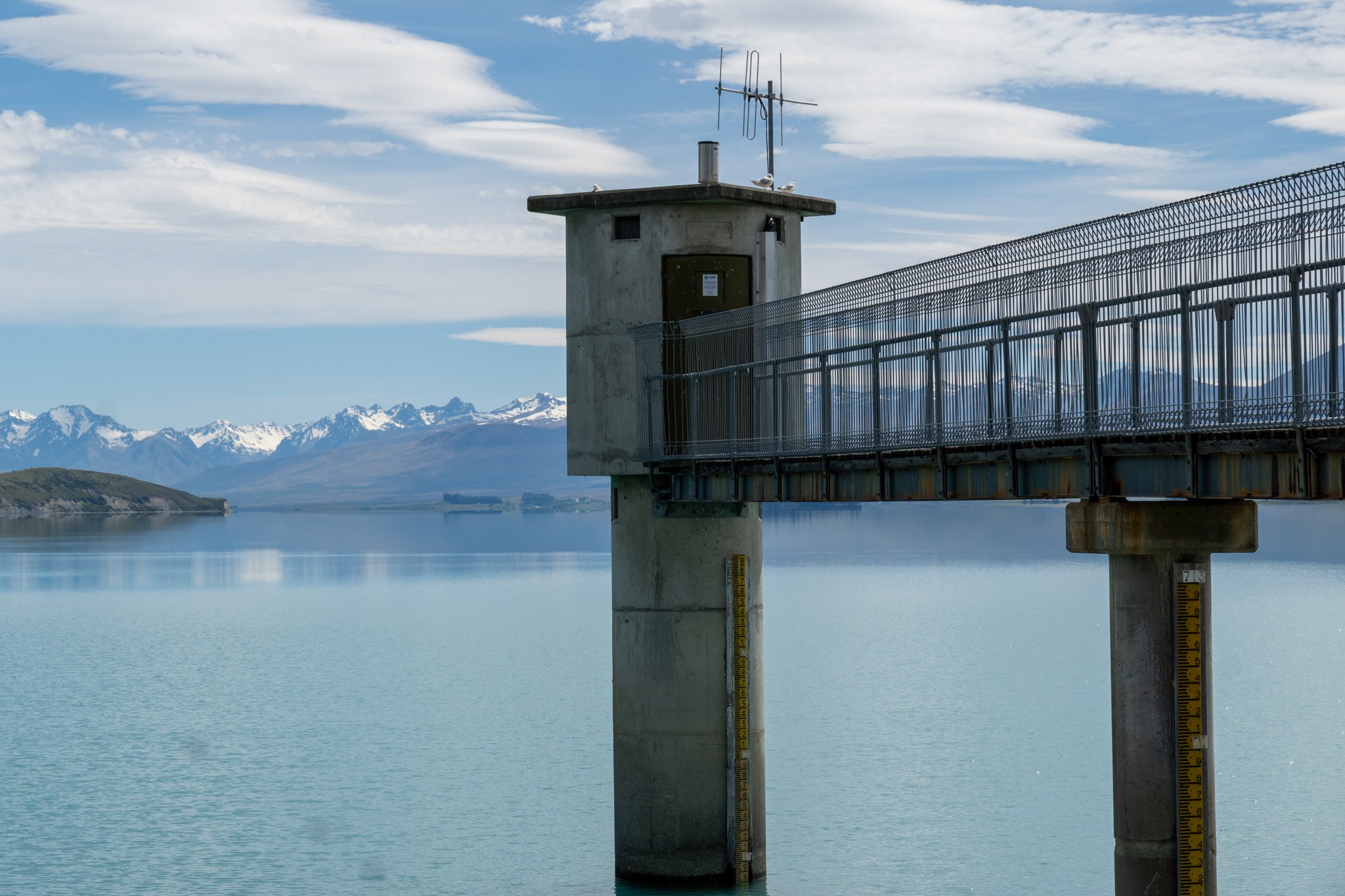 A concrete water level gauge attached to a pier over a body of water, with a small control building on the pier, in a landscape with snow-capped mountains, blue sky, and clouds in the background.