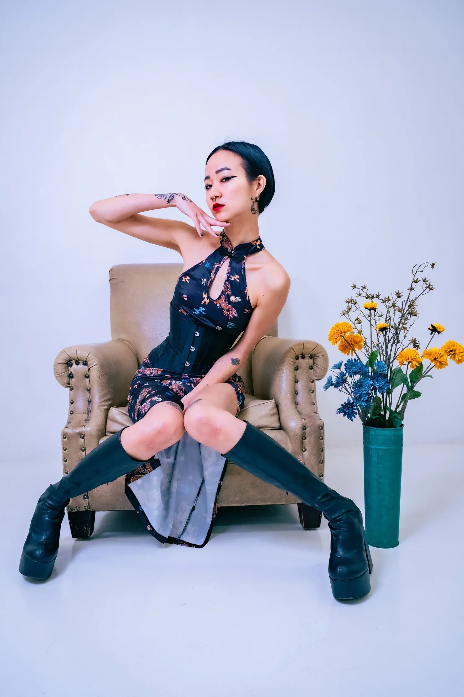 Mistress Koto with black, wavy hair in a stylish outfit and tall black boots standing next to a motion capture camera on a wooden floor with dark curtains in the background.