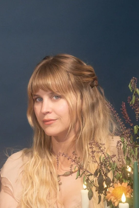 A woman with long blonde hair and bangs, wearing a light-colored top, posing in front of a dark blue background with floral decorations and candles.