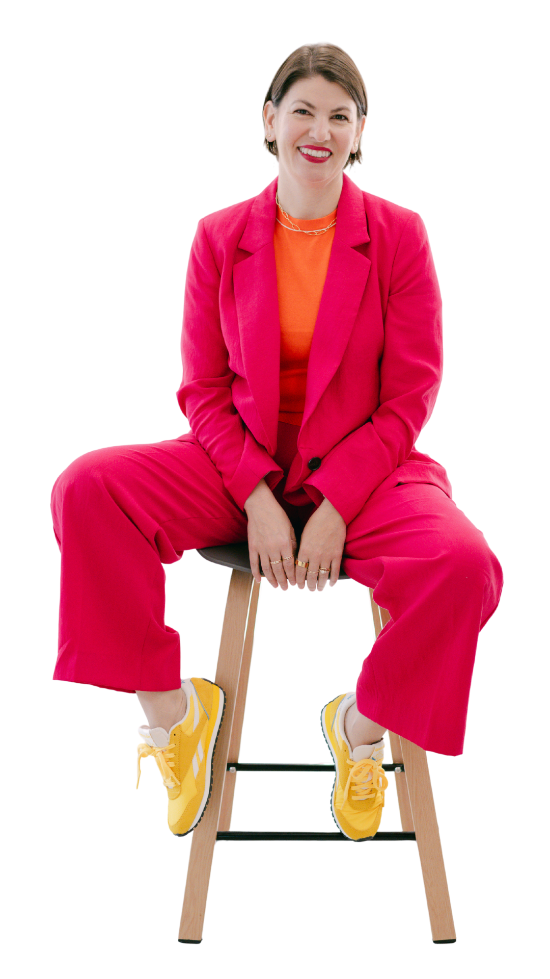 A woman is sitting on a wooden stool, smiling, wearing a bright pink suit with an orange top, yellow sneakers, and some jewelry.