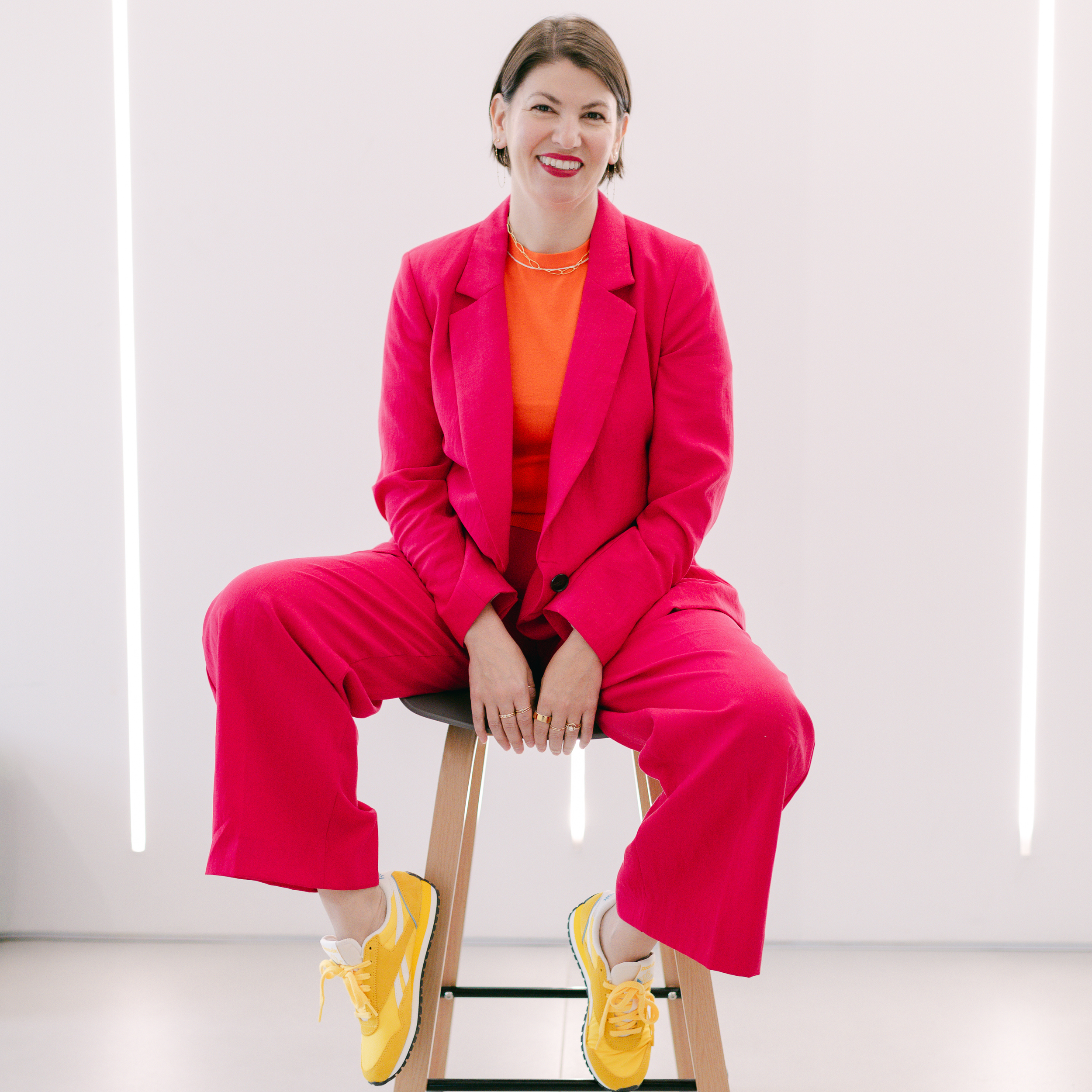Woman sitting on a stool against a bright background, wearing a pink suit, orange top, and yellow sneakers, smiling at the camera.
