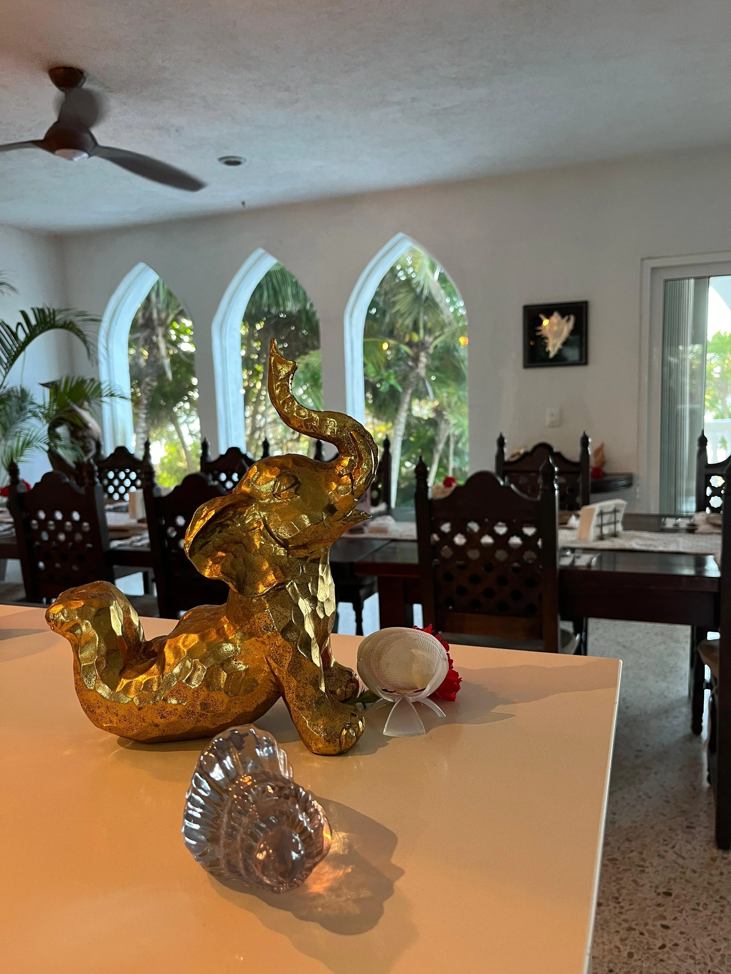 Golden decorative sculpture resembling a mythical creature, placed on a table with small seashells and red flowers, in a room with arched windows, framed artwork on the wall, potted plant, and dining chairs in the background.