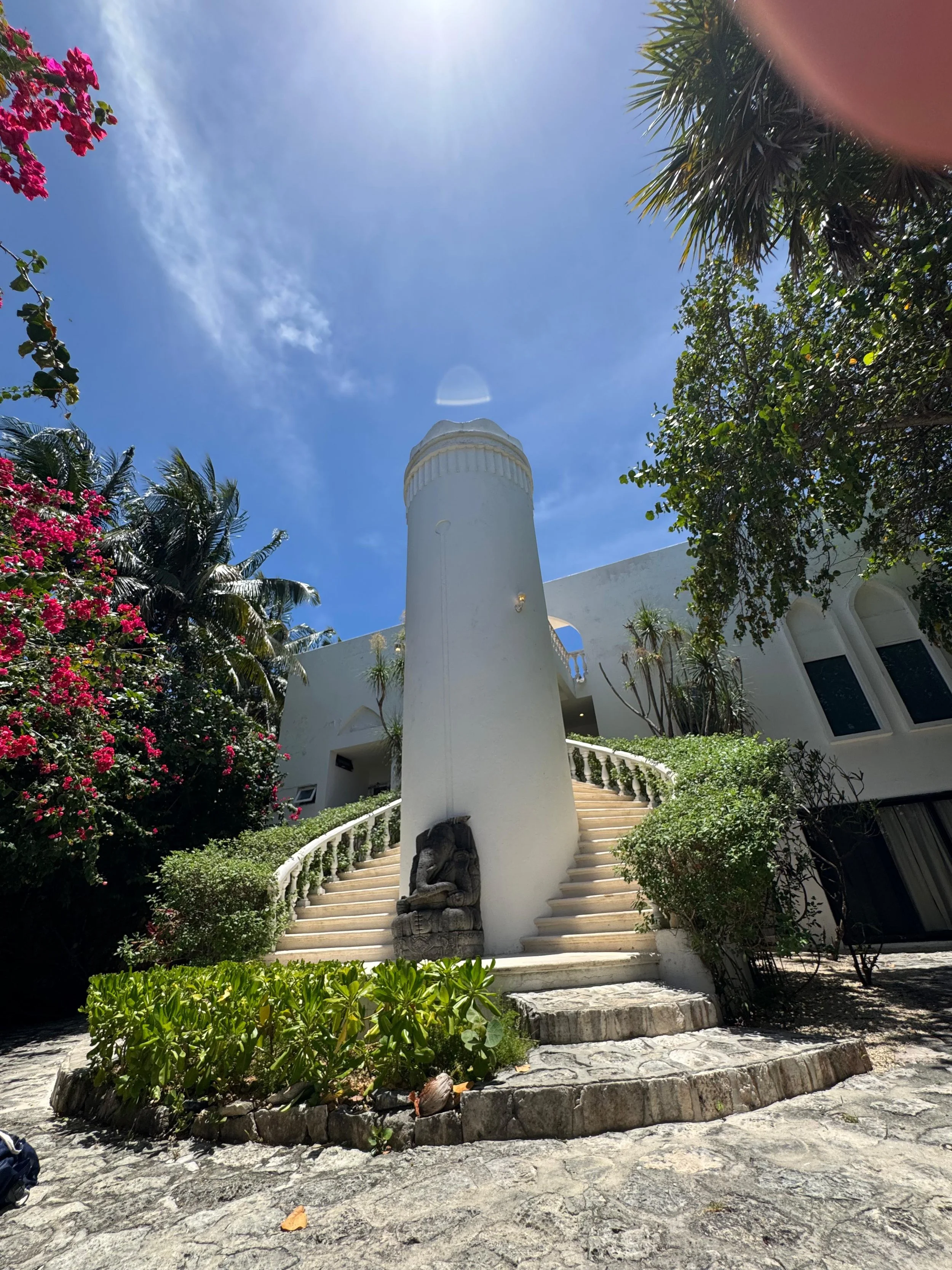 A tall white building with curved stairs and a sculpture of an elephant at the base, surrounded by colorful flowers and tropical plants under a bright blue sky.