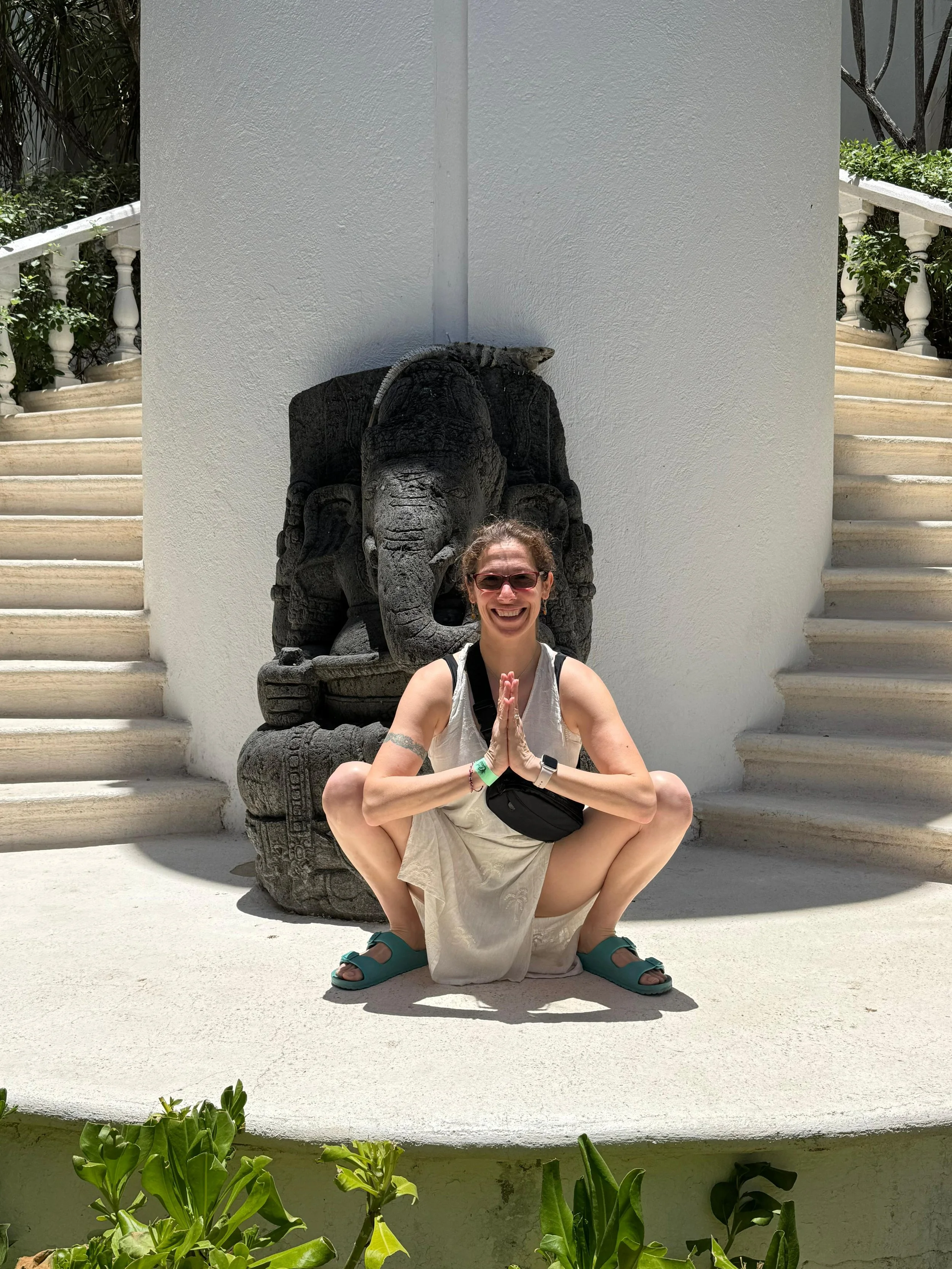 A woman with short curly hair, wearing sunglasses, a white tank top, a wristwatch, and green sandals, is doing a yoga pose in front of a stone elephant sculpture at a staircase landing, with white walls, greenery, and stairs in the background.