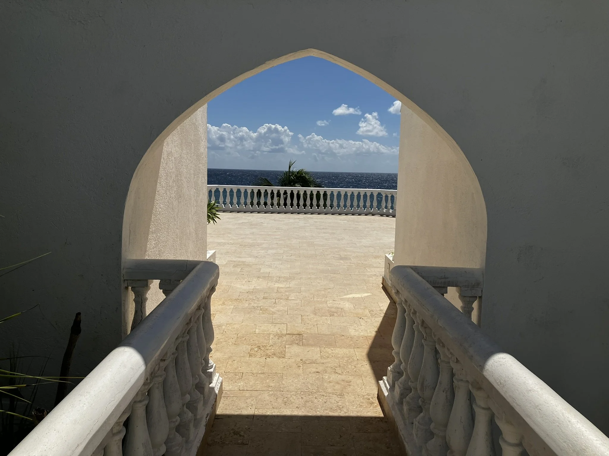 View of the ocean with a blue sky and white clouds seen through an archway, with a white balcony and palm trees in the foreground.