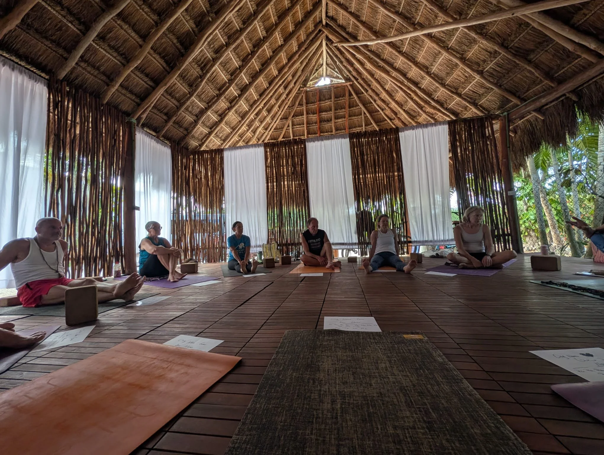 People participating in a yoga or meditation class inside a rustic, open-air structure with a thatched roof, wooden support beams, and white curtains.