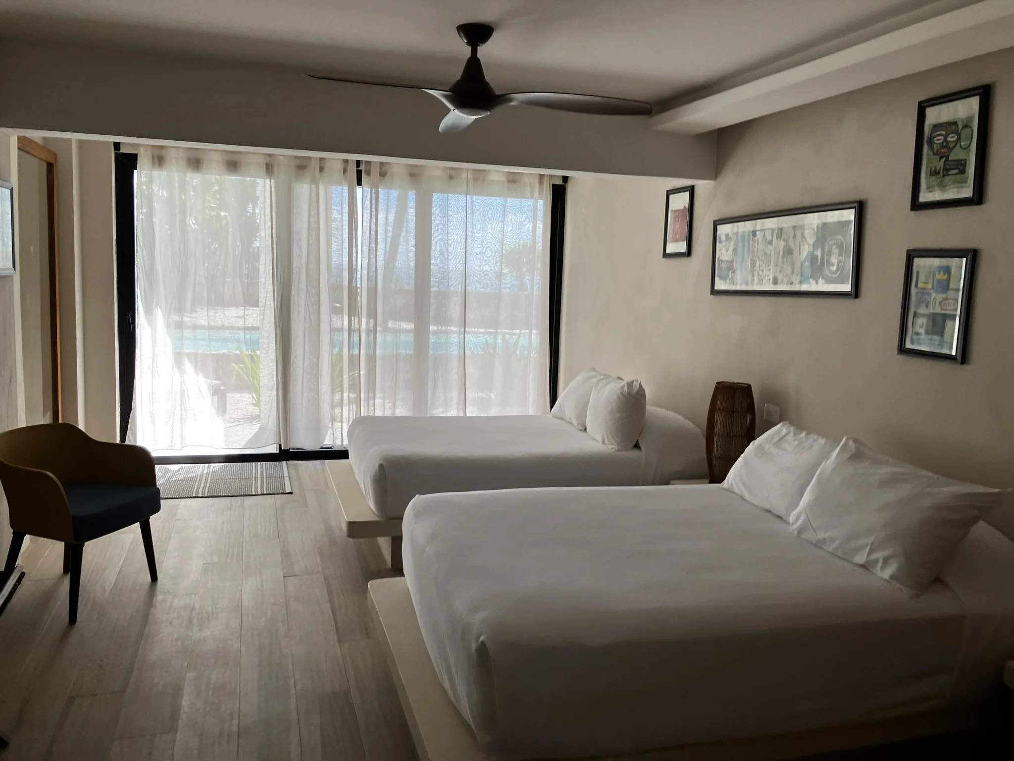 A living room with large glass doors covered by sheer curtains, a ceiling fan, framed artwork on the wall, and white sofas with pillows.