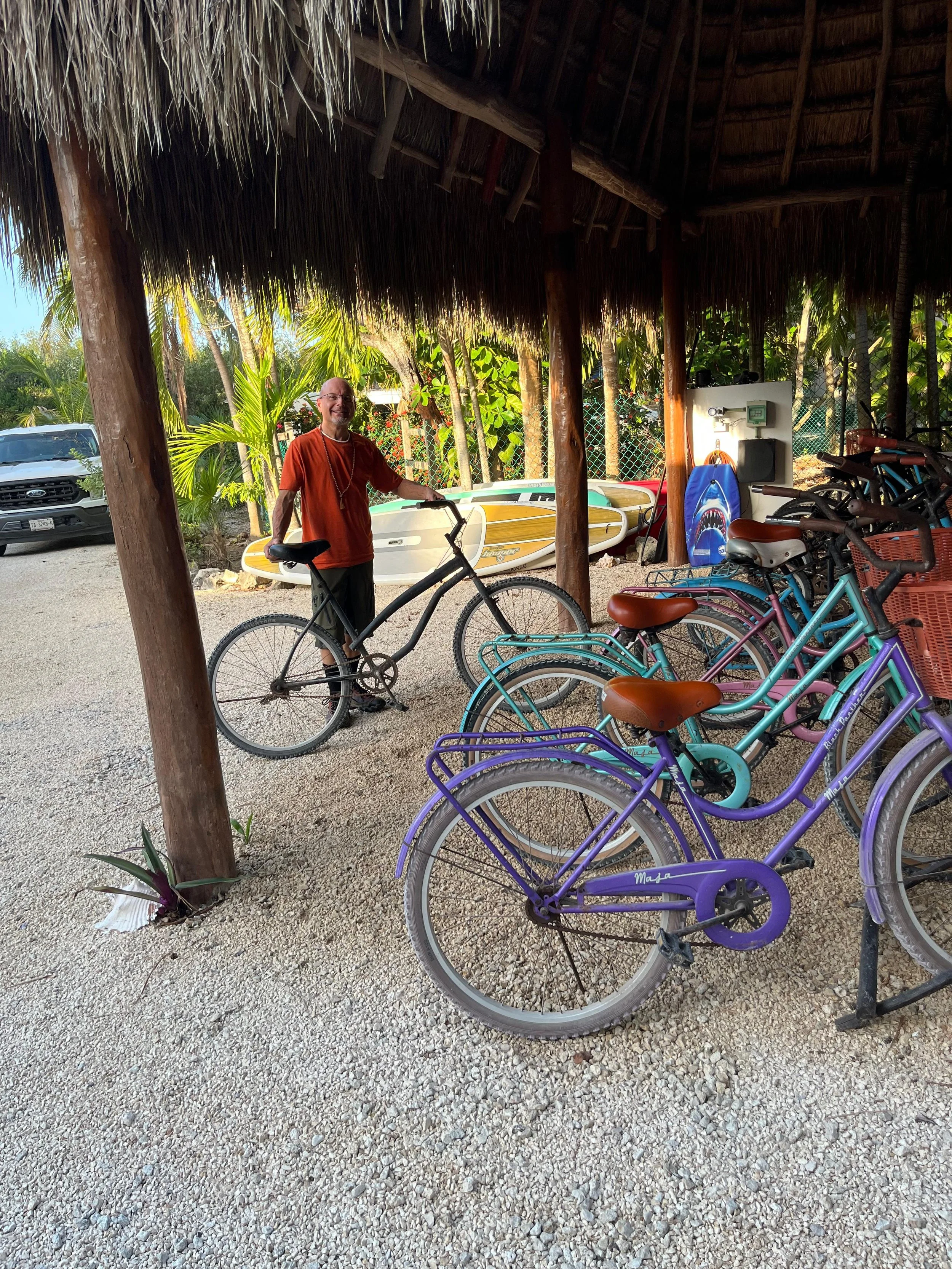 A man smiling and holding a black bicycle in a shaded tropical area with colorful bikes, paddleboards, and lush green foliage in the background.