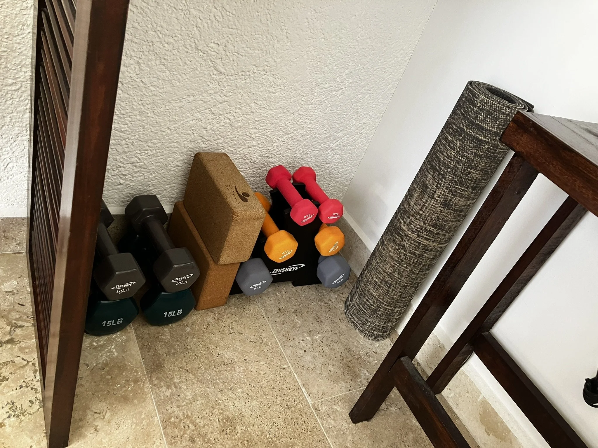 A collection of exercise equipment including dumbbells, yoga mats, a cork yoga block, and a foam roller, stored on the floor next to a wooden table and a white wall.