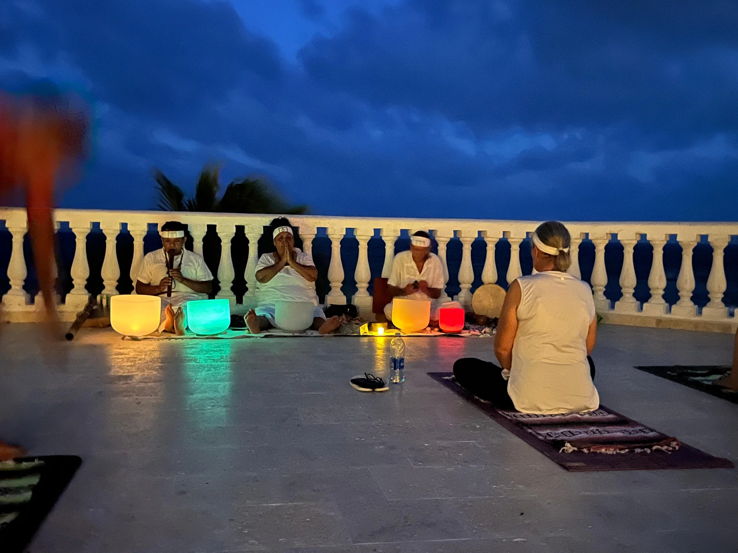 People sitting on the floor in front of a white balustrade, participating in a spiritual or meditation session outdoors during dusk, illuminated by colorful lamps, with a dark sky and a palm tree in the background.