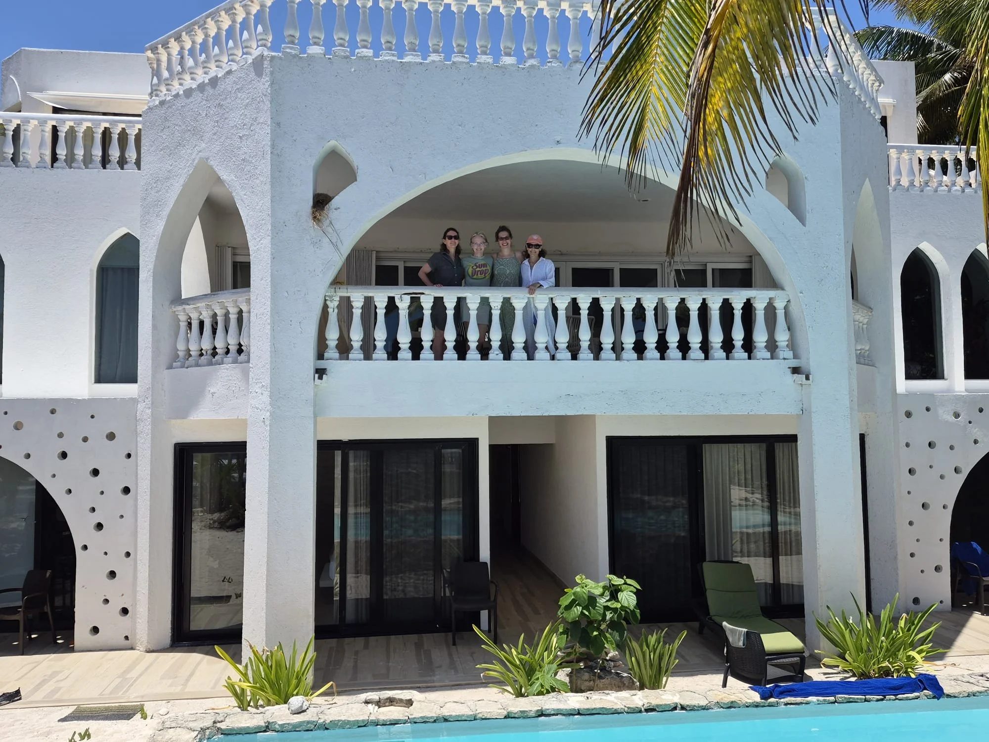 A white building with arched openings, a terrace with a railing, and tropical palm trees. Four women stand on the balcony beneath the arch, with lush greenery and a swimming pool in the foreground.