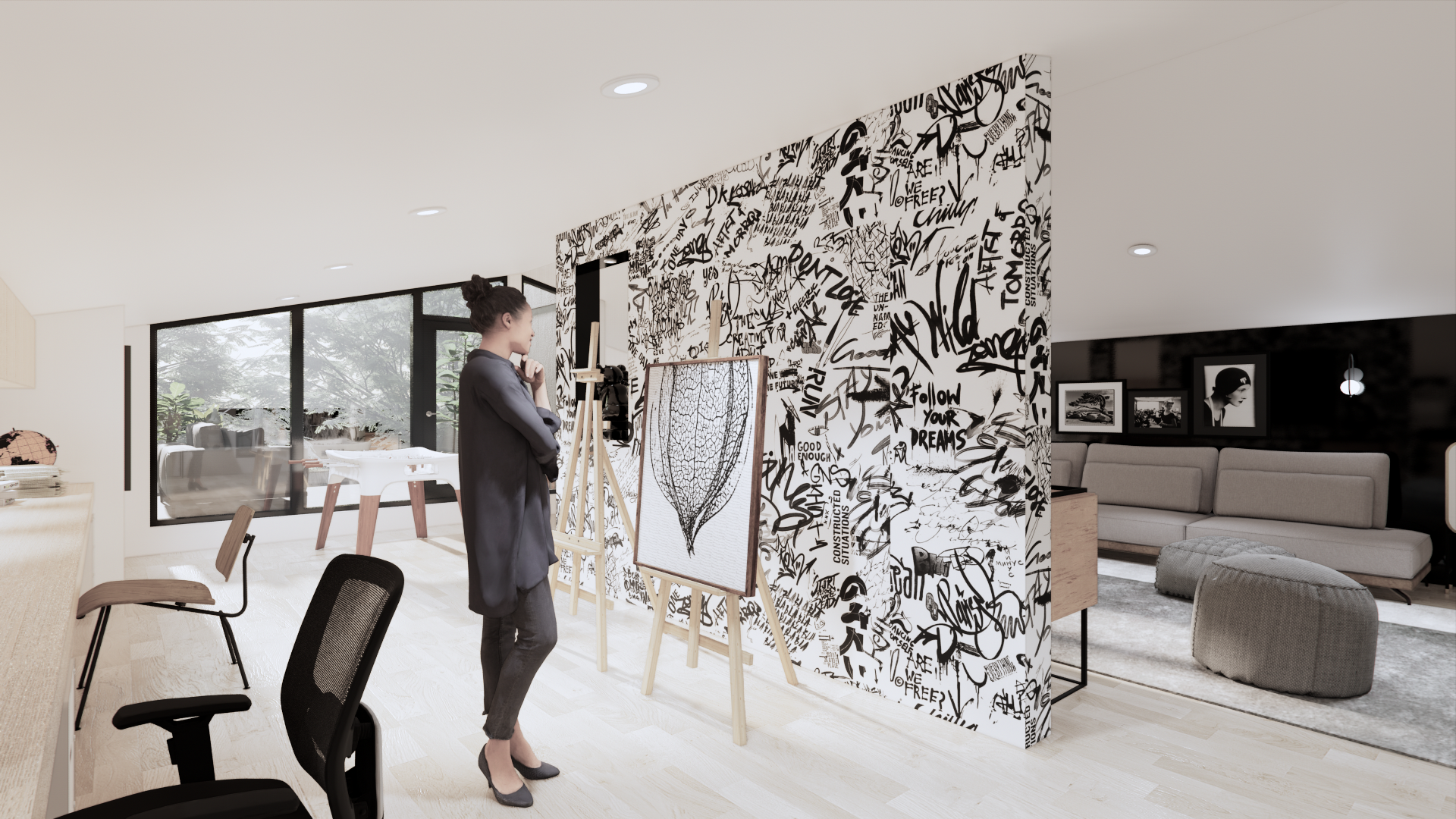 A woman standing in an art studio, observing a drawing on an easel. The studio has a black and white graffiti-style wall, large windows with a view of trees, and a seating area with a gray couch and ottomans.
