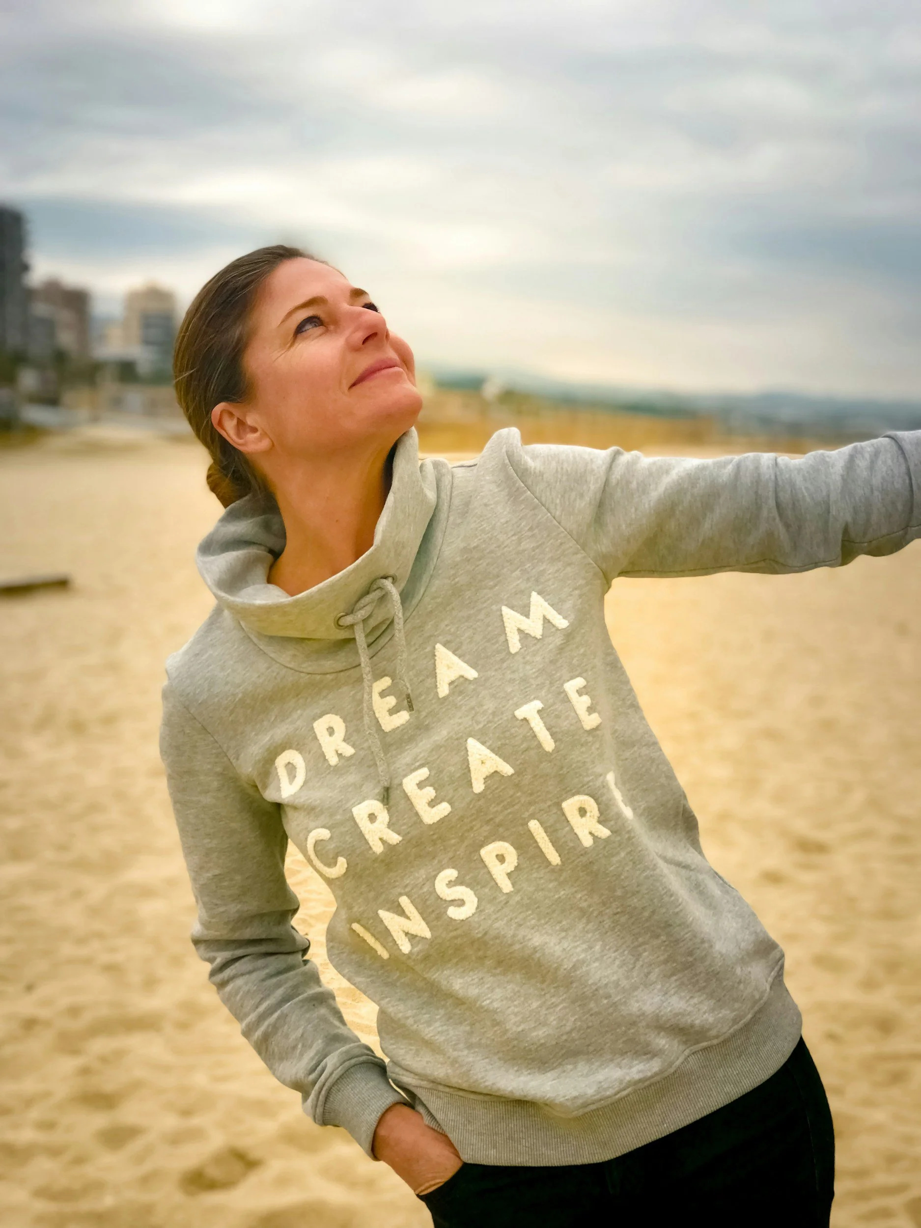 A woman standing on a sandy beach, smiling and looking upward, wearing a gray hoodie with the words 'DREAM CREATE INSPIRE' printed on it, with an urban skyline blurred in the background.