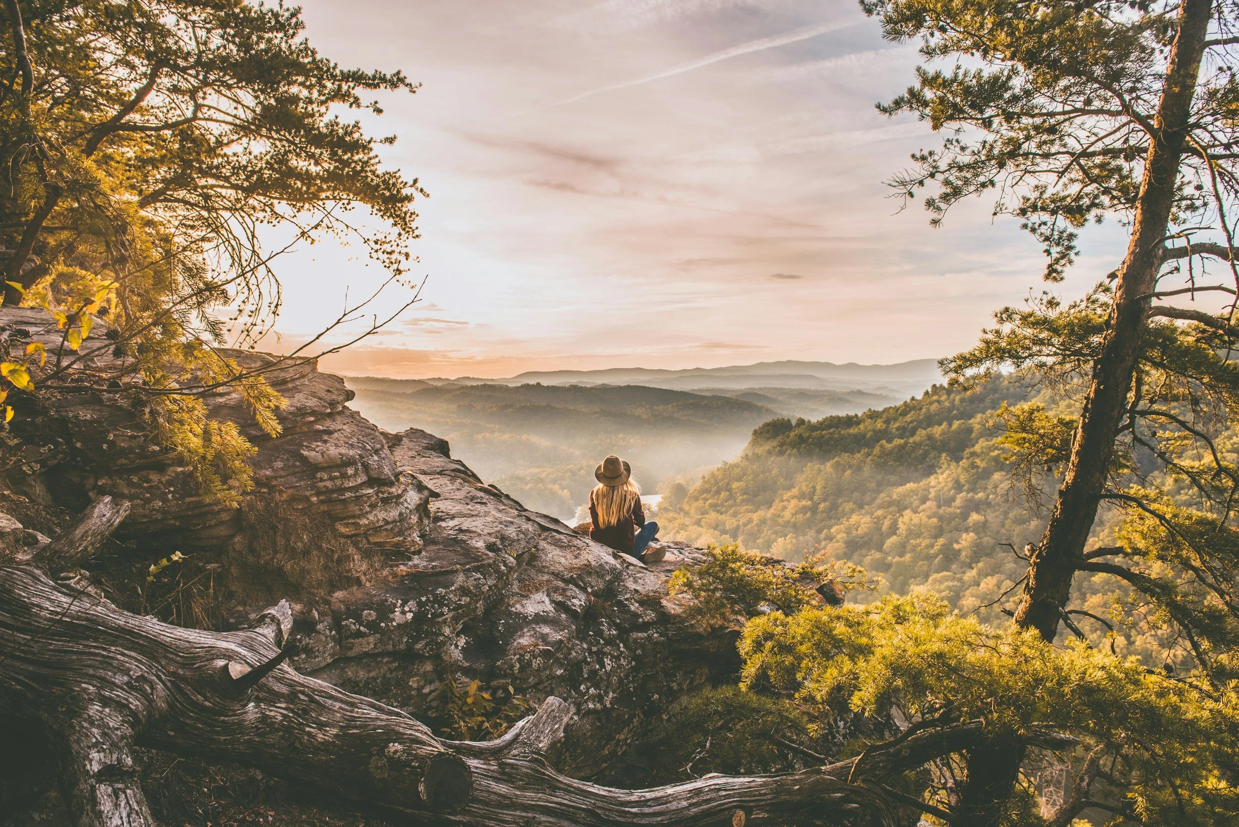 A person sitting on a rocky cliff overlooking a forested valley during sunset, wearing a hat and casual clothing.
