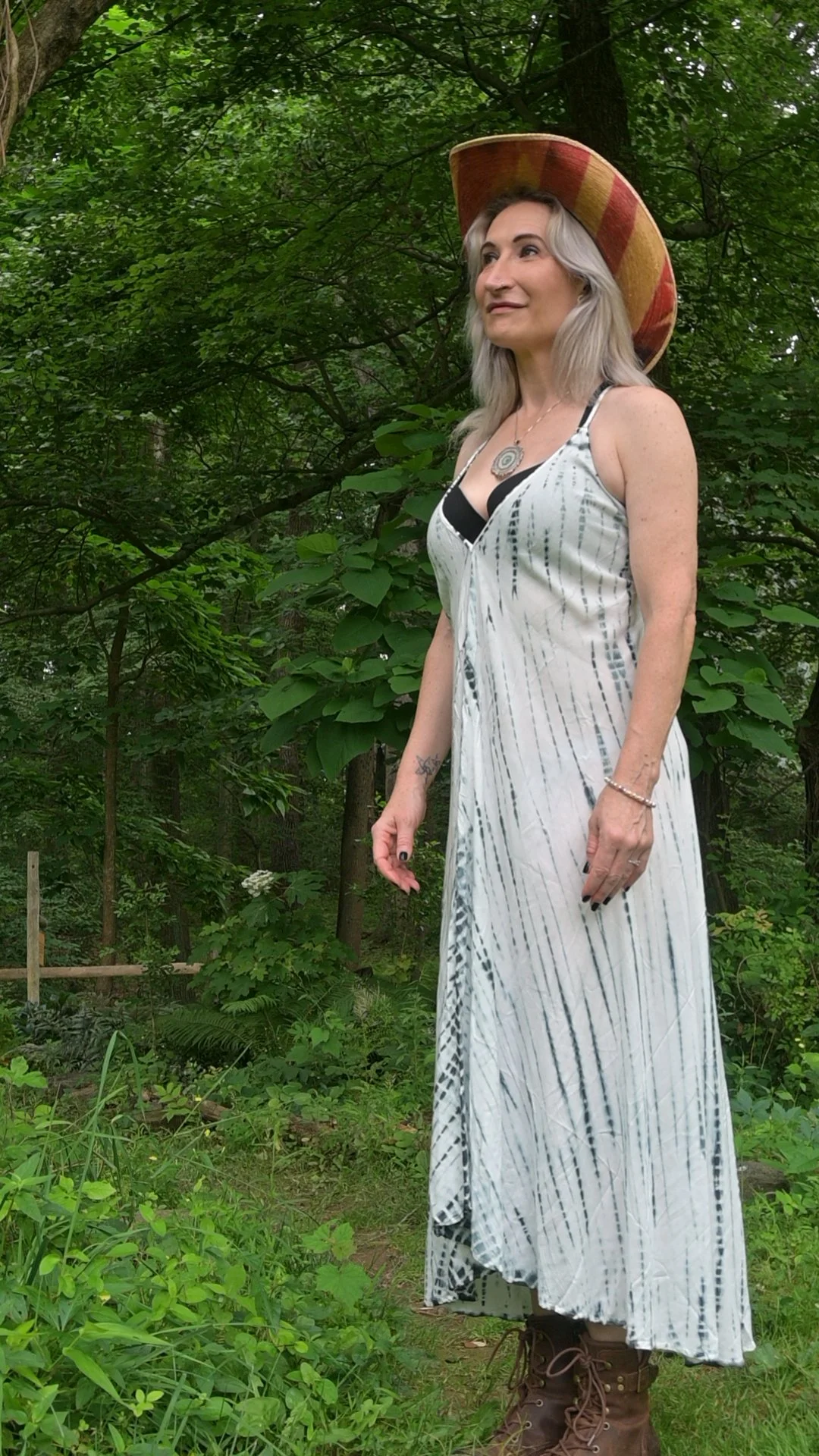Woman standing outdoors in a forested area, wearing a long, flowy, patterned dress, a large striped hat, and brown lace-up boots.
