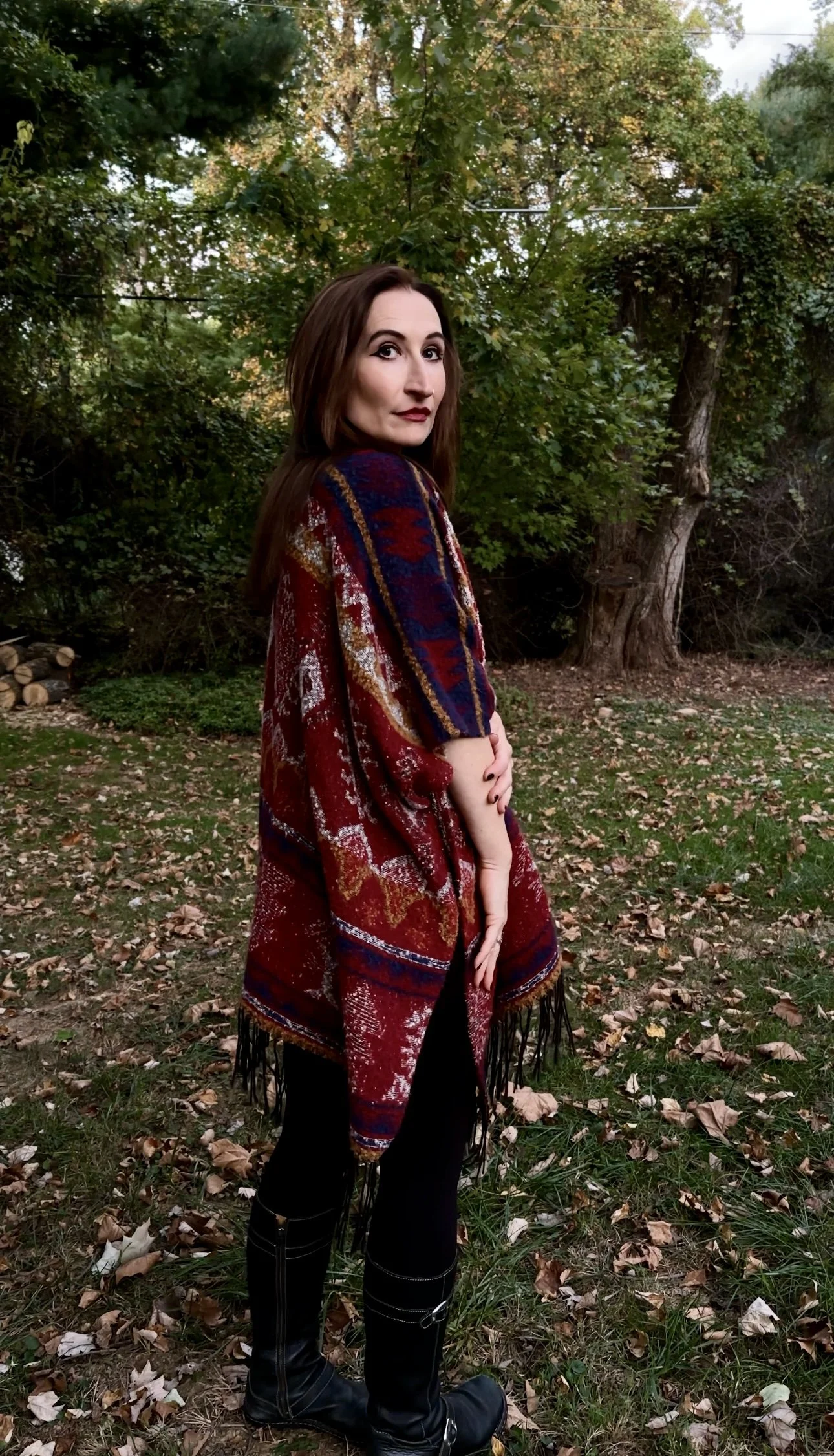 A woman with long brown hair standing outdoors in a wooded area with fallen leaves, wearing a multicolored patterned shawl, black pants, and knee-high black boots, looking back at the camera.