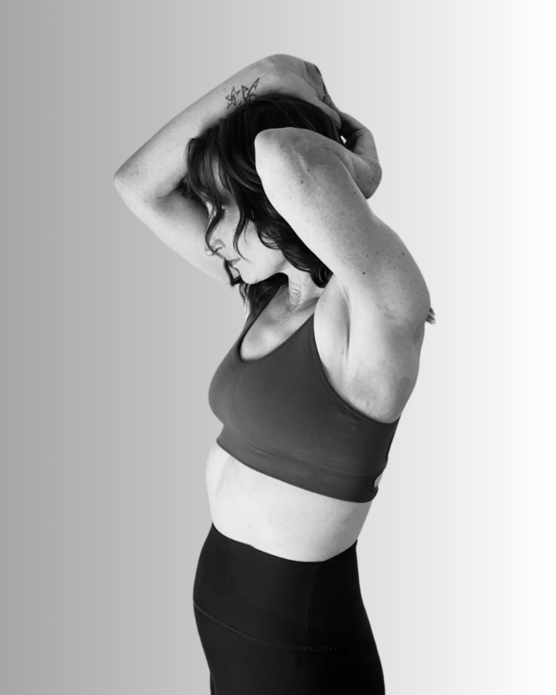 Black and white photo of a woman with dark hair in a sports bra and high-waisted leggings, standing with arms raised, hands behind her head, in a contemplative pose.