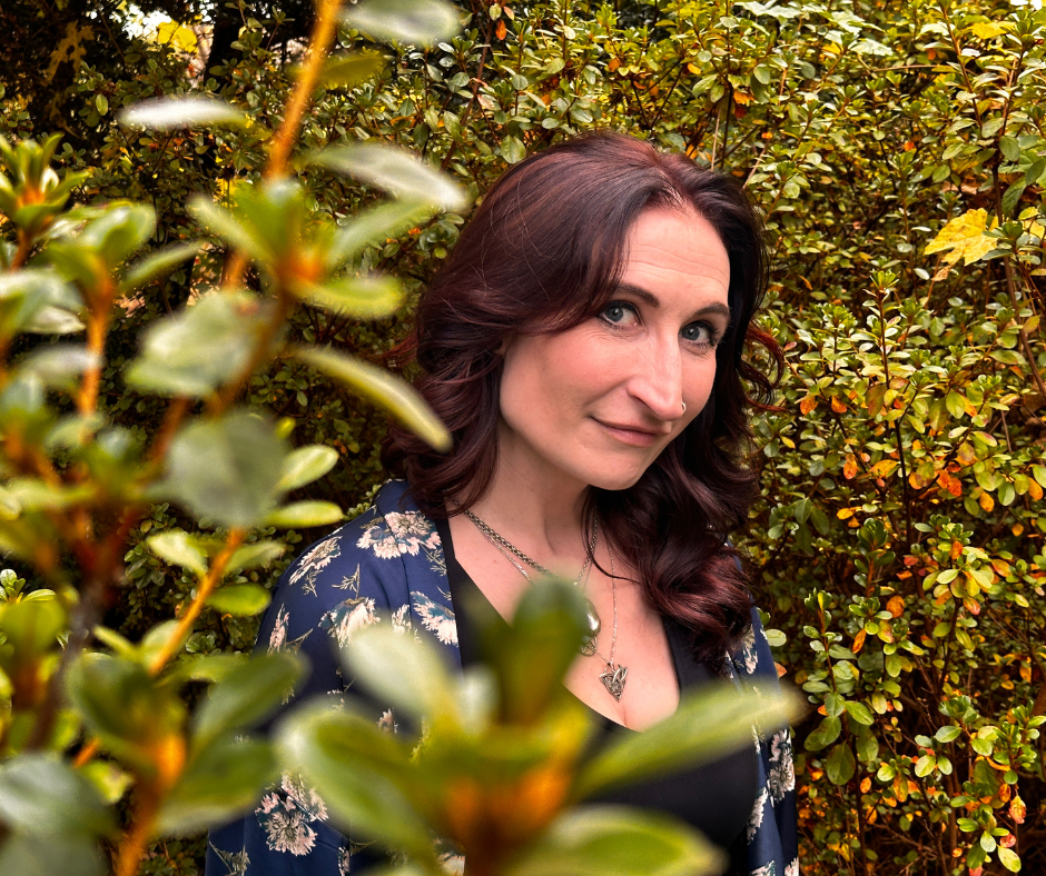 A woman with dark hair and blue eyes standing among green and yellow foliage, wearing a floral kimono-style jacket and a heart-shaped necklace, looking at the camera with a slight smile.