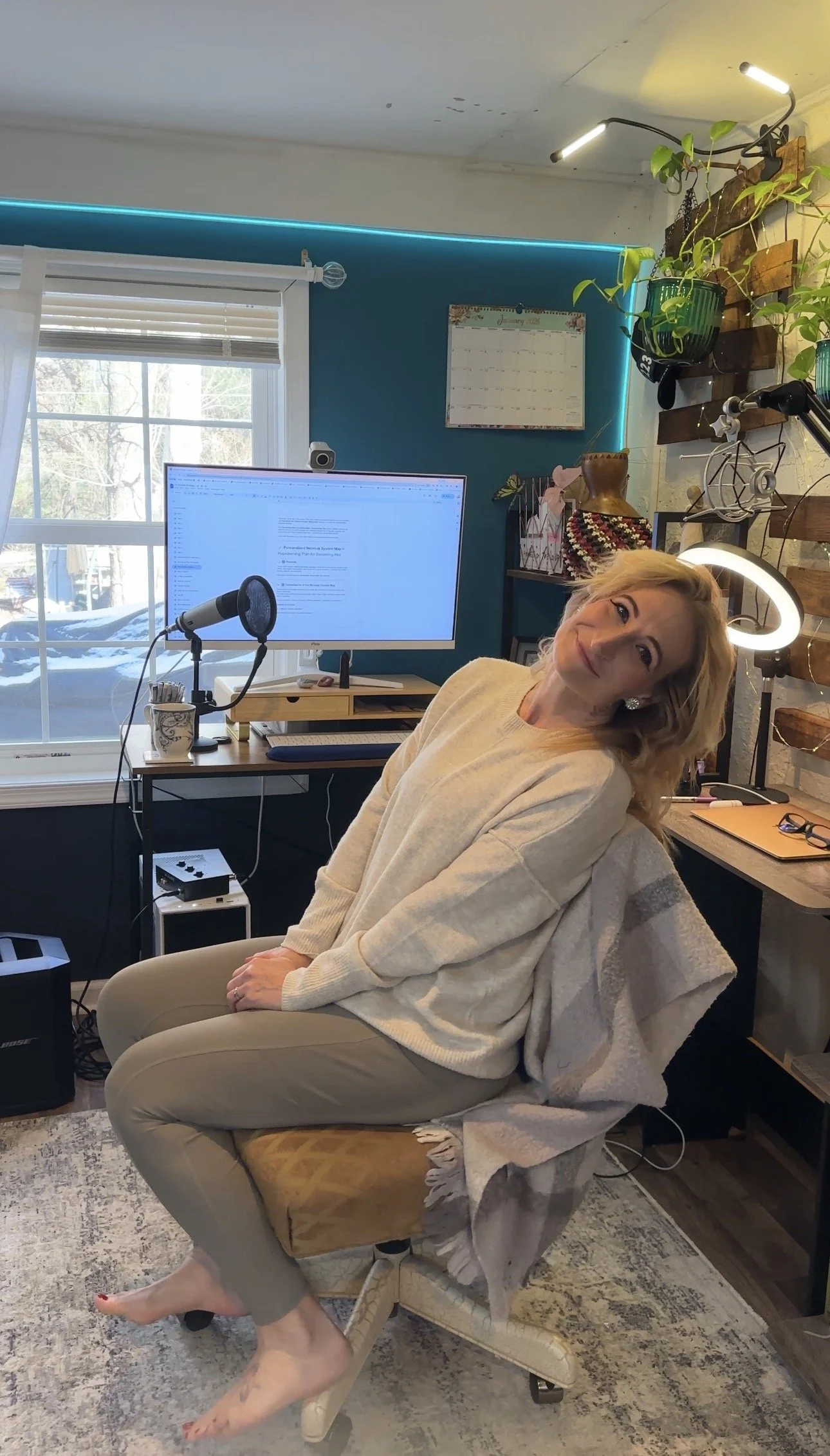 A woman sitting on a beige office chair with a white throw blanket, in a home office with a large window, a computer monitor, a microphone, and green plants.