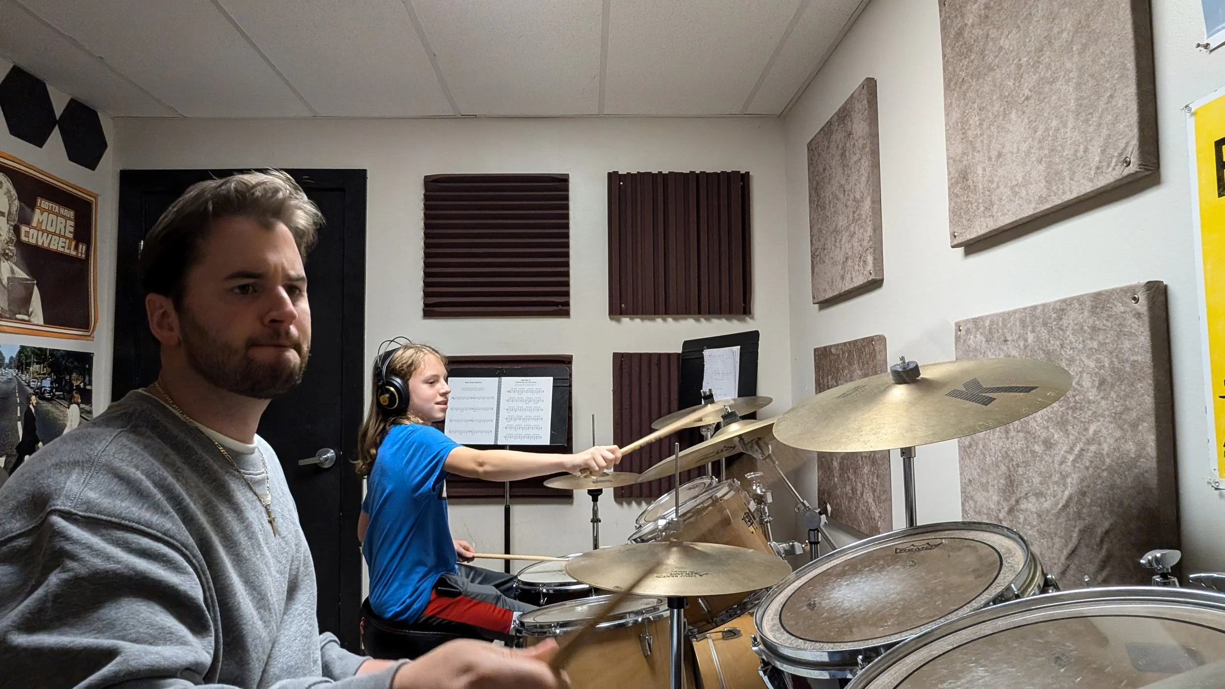 Two people in a music recording studio. The man on the left is sitting at a drum set, and the young girl in the background is playing the drums with drumsticks, wearing headphones.