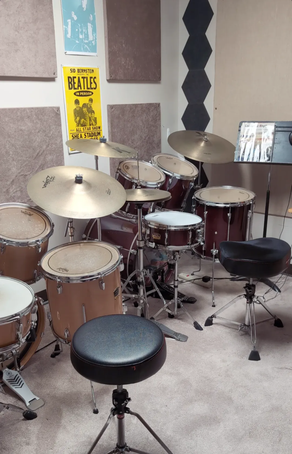 A drum set with various drums and cymbals in a music practice room, with acoustic panels on the wall and a yellow Beatles concert poster.