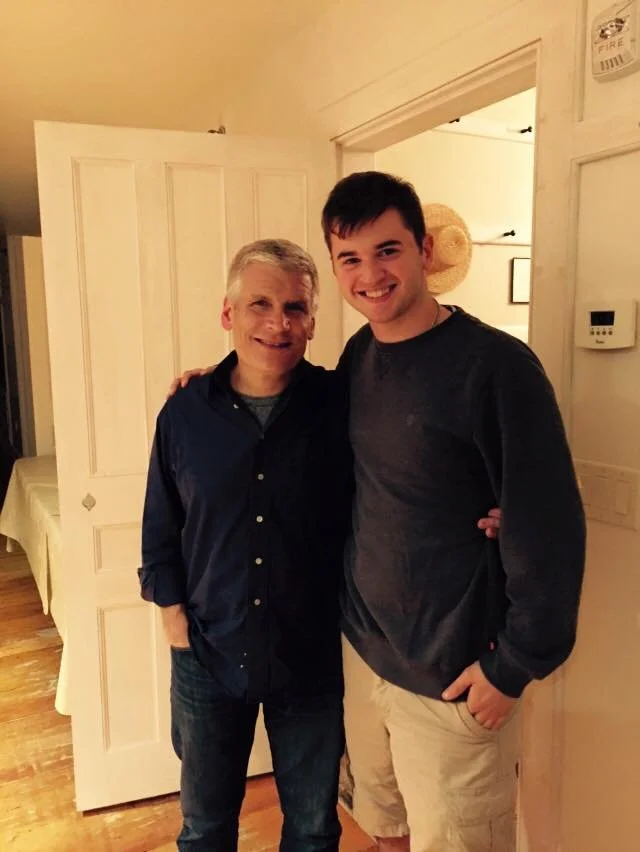 Two men standing indoors, smiling and embracing, with a doorway behind them. One has short gray hair and is wearing a navy shirt, the other is younger with dark hair and a black shirt.