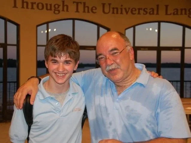 A young boy and an older man are smiling and standing with their arms around each other inside a building with large arched windows and a view of a body of water outside. The background has a banner with the text "through The Universal Lang".