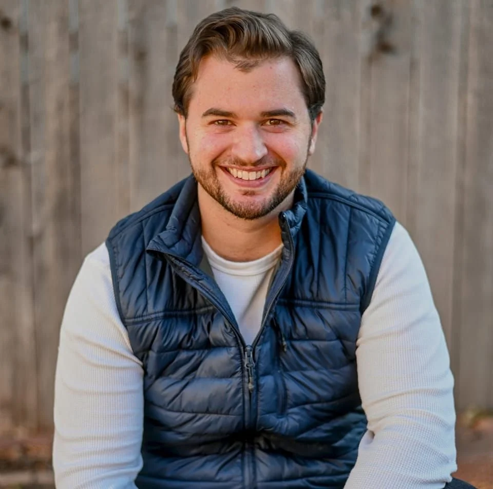 A young man with brown hair and a beard smiling, wearing a white shirt and a navy blue quilted vest, sitting in front of a wooden fence.