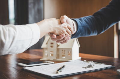 Two people shaking hands over a table with a small house model, an open notebook, and a pen.