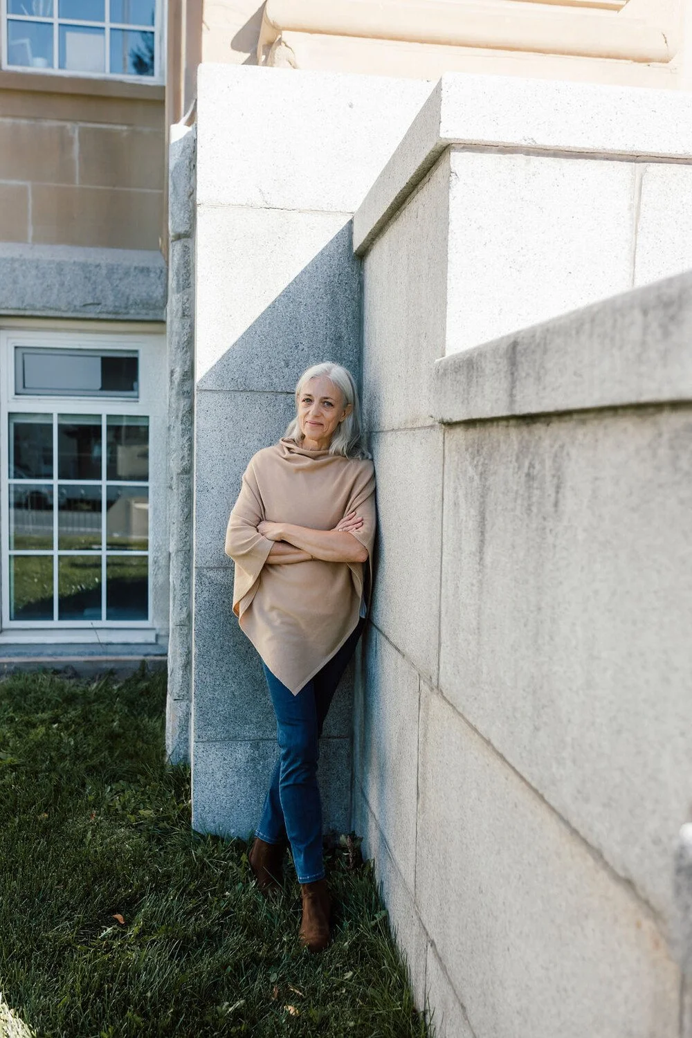 Angelique de Pennart leaning against a stone wall outside a building, wearing a beige poncho, blue jeans, and brown boots.