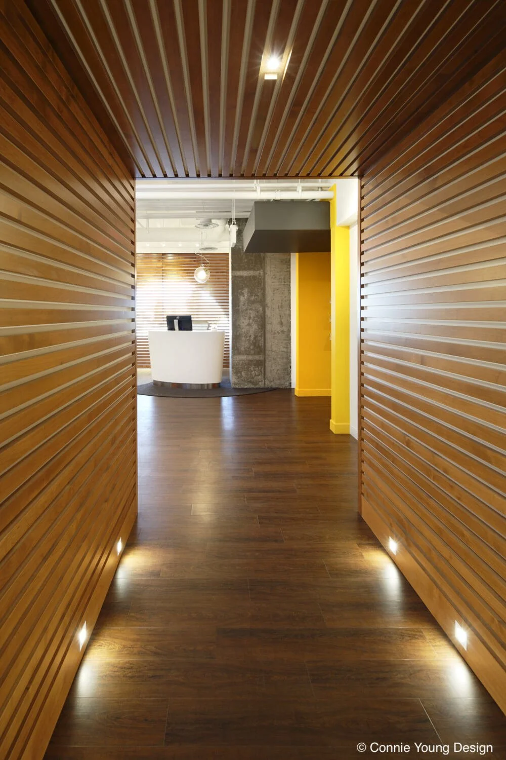 Modern office reception area with white curved desks, computer monitors, a glass display shelf with jars, wooden blinds, and a concrete pillar.