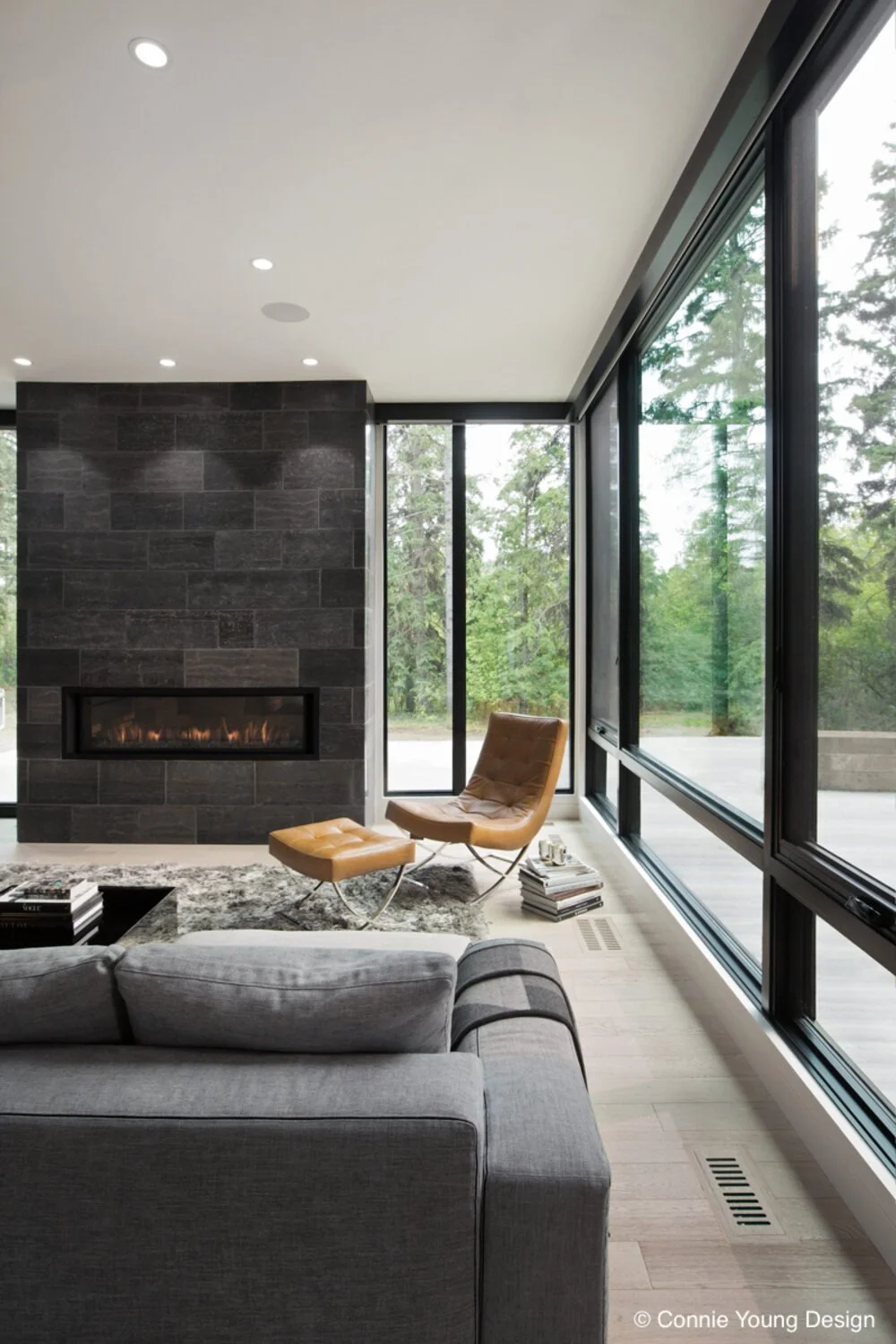 Living room with large glass windows showcasing a wooded outdoor scene, a modern fireplace, a tan lounge chair with matching ottoman, a gray sofa, a stack of books, and a textured area rug.