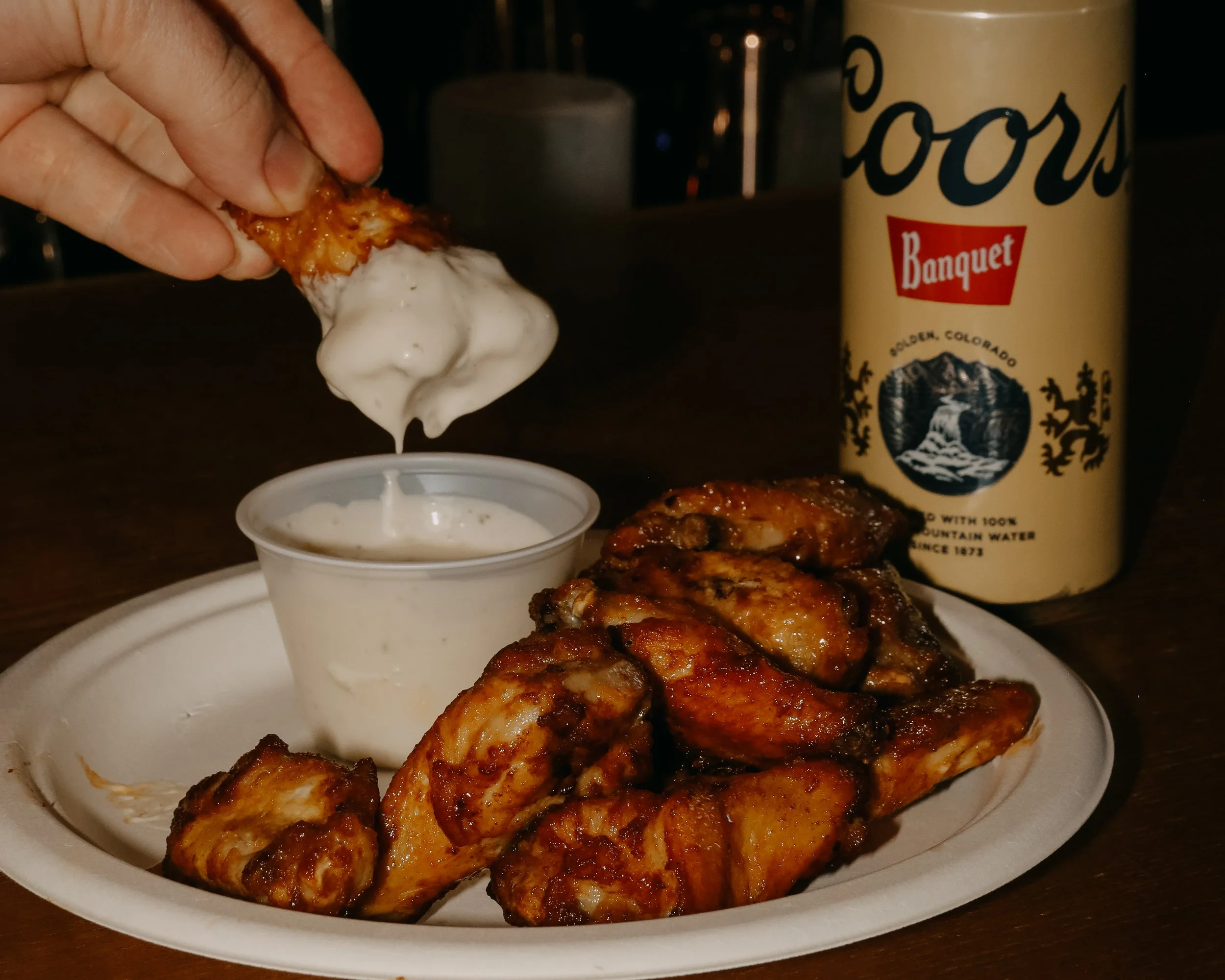A hand dipping a chicken wing into a cup of ranch dipping sauce, with a plate of fried chicken wings and a cup of sauce, and a Coors Banquet beer can in the background.