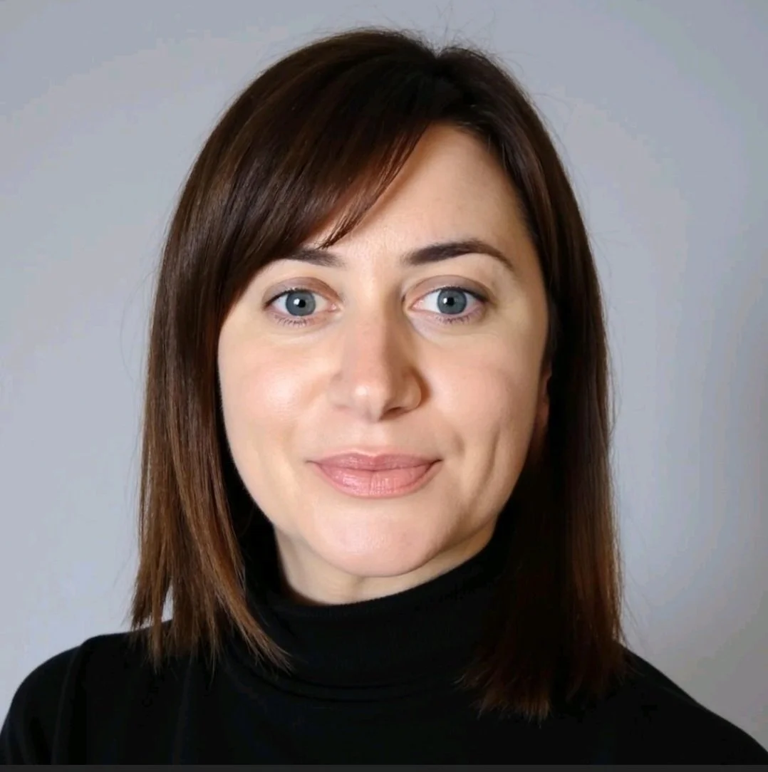 Close-up portrait of a woman with shoulder-length brown hair and blue eyes, wearing a black turtleneck, smiling softly against a gray background.