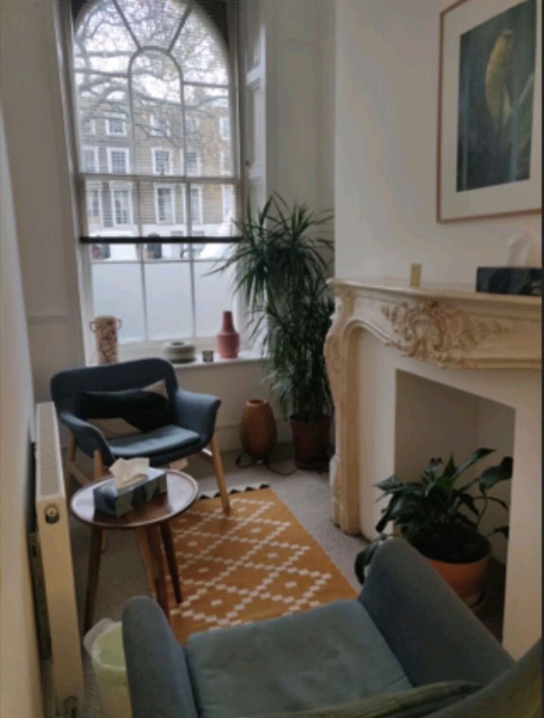 A cozy living room with a large window, a blue armchair, a small round wooden table with tissue box, indoor plants, decorative vases, a fireplace, and wall art